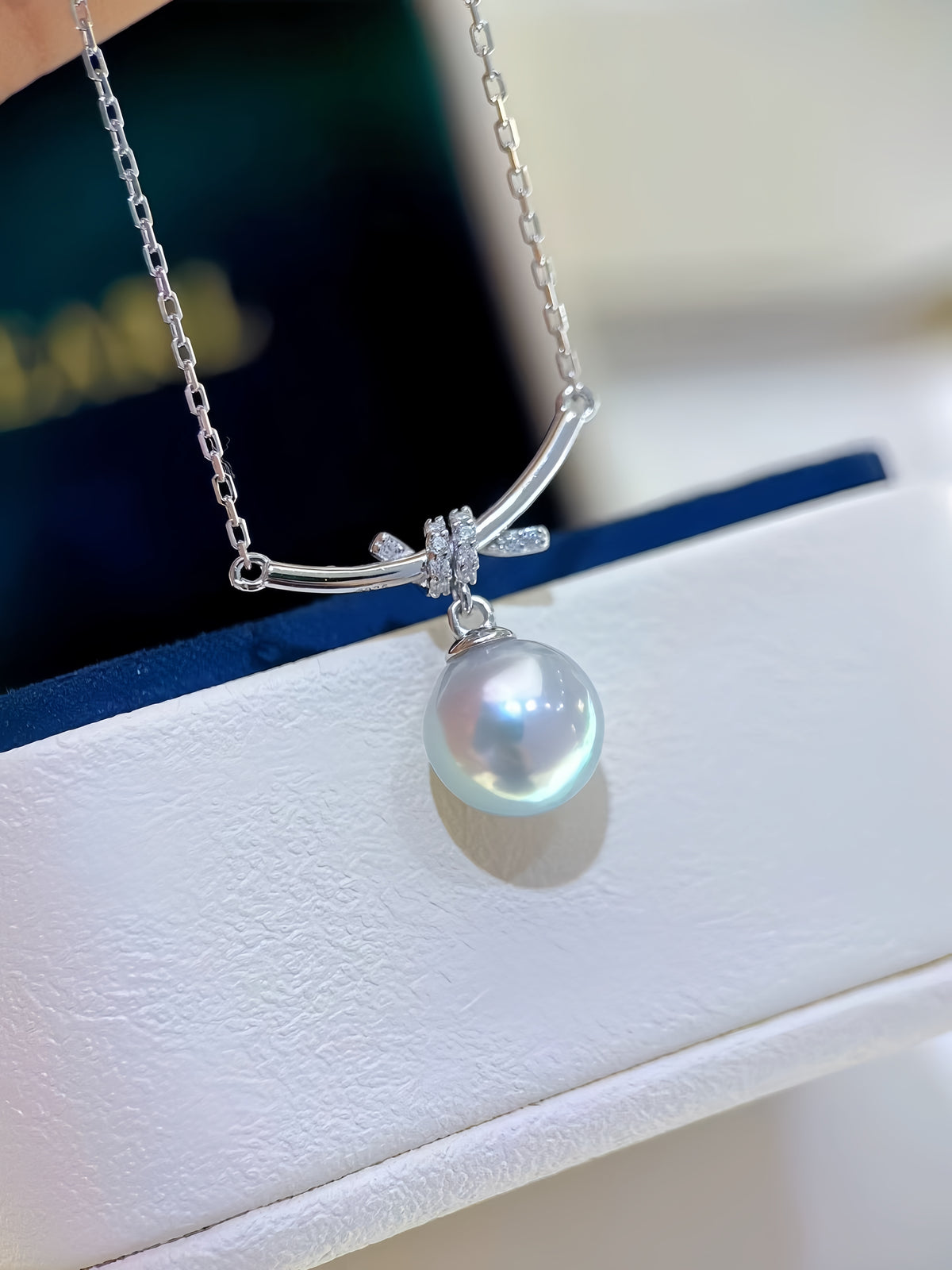 A close-up of a silver-tone necklace with a curved bar, a small bow-shaped cluster of clear stones, and a single lustrous pearl pendant, shown against a blurred dark blue background.