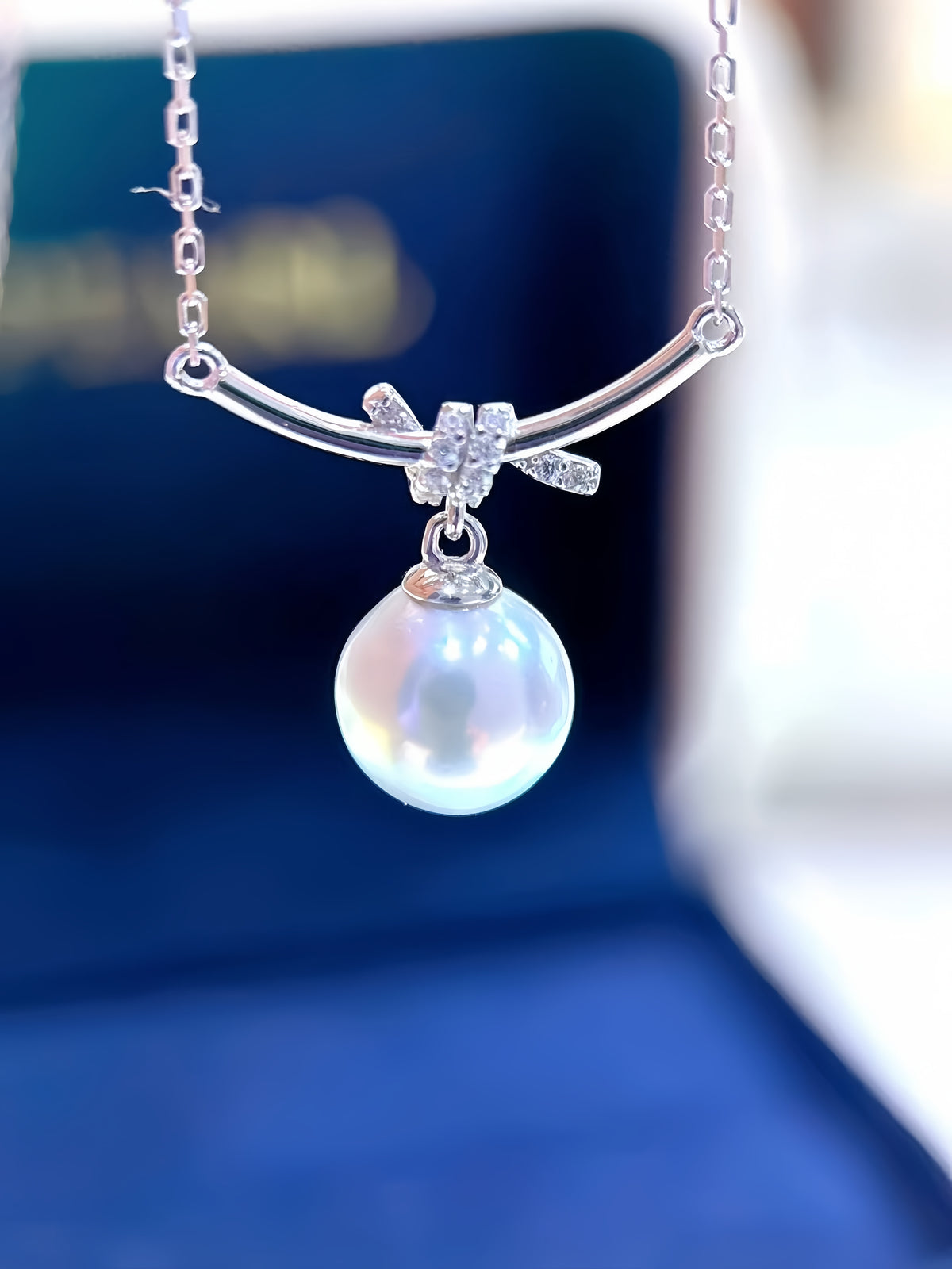 A close-up of a silver-tone necklace with a curved bar, a small bow-shaped cluster of clear stones, and a single lustrous pearl pendant, shown against a blurred dark blue background.