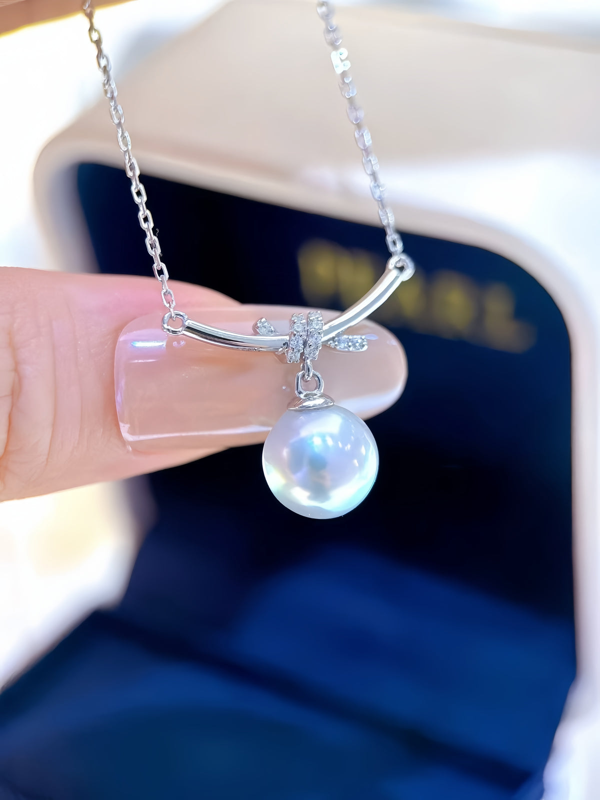 A close-up of a hand holding a silver necklace with a curved bar, small clear stones, and a single lustrous pearl pendant. The necklace is positioned in front of an open dark blue jewelry box.