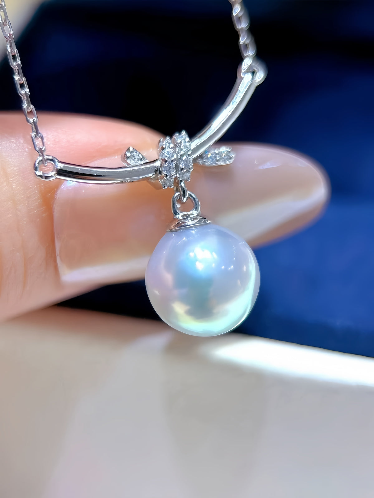 A close-up of a silver necklace with a curved bar pendant, a small floral cluster of clear stones, and a large round iridescent pearl pendant, held between fingers against a dark blue background.