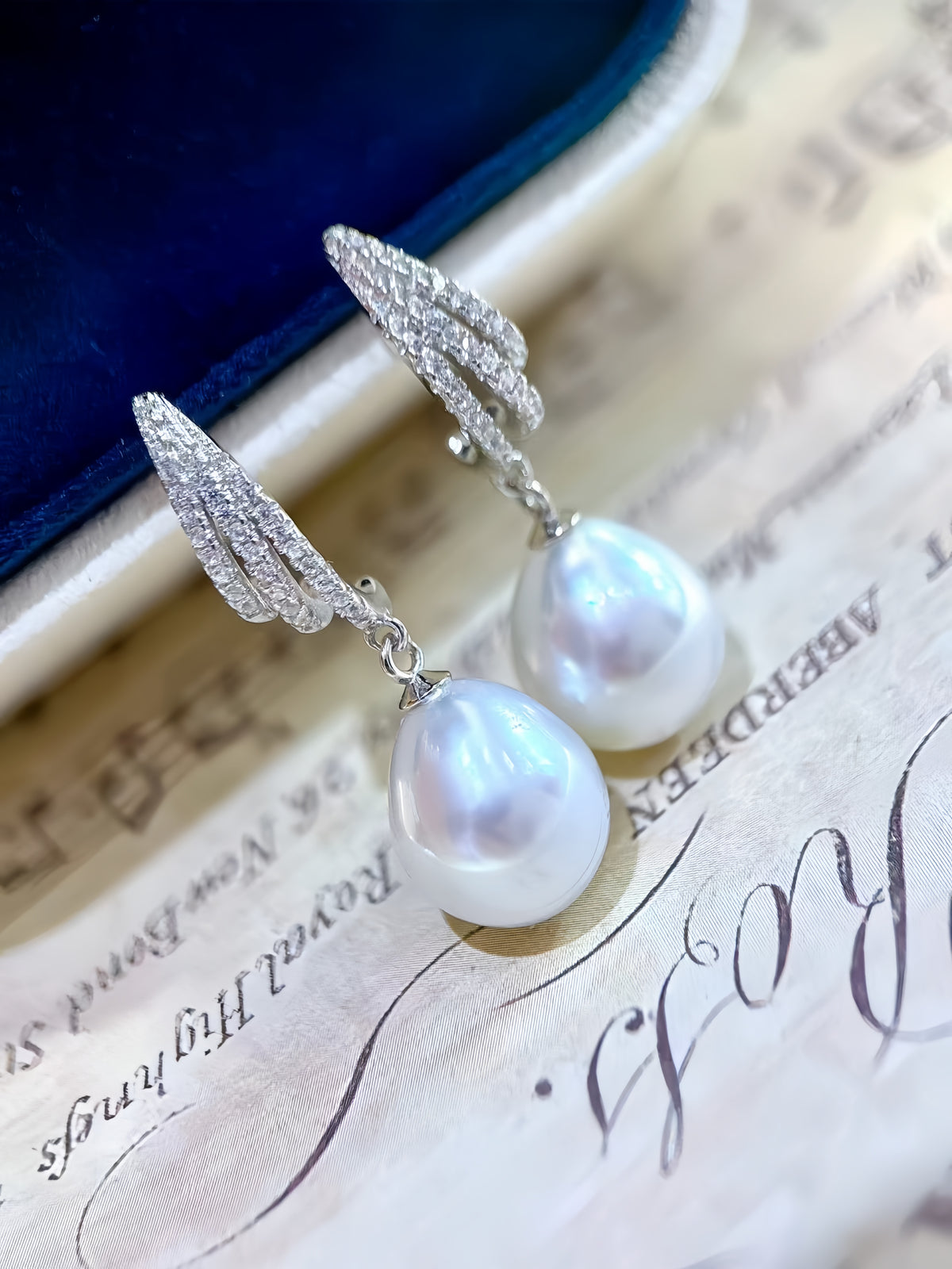 A pair of silver drop earrings with teardrop-shaped white freshwater pearls and sparkling clear stones in a spiral design, resting on a vintage-style paper background with a dark blue velvet surface.