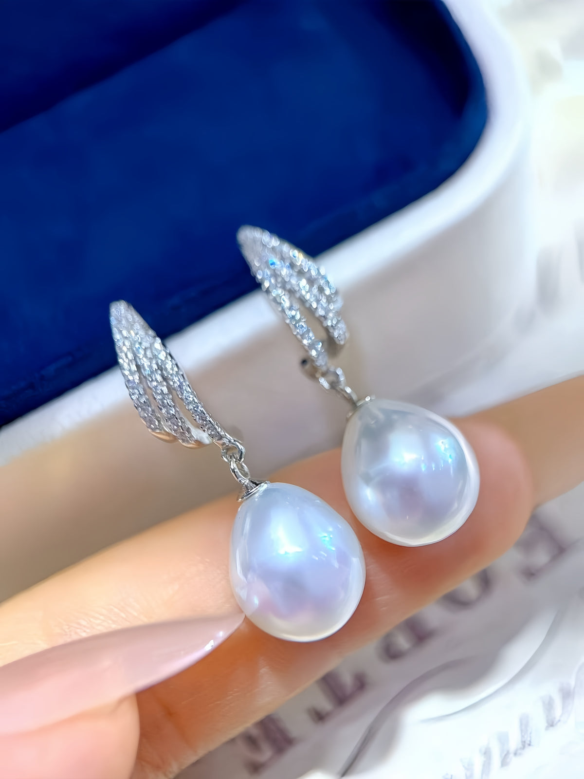 A pair of silver huggie hoop earrings with teardrop-shaped white freshwater pearls, held between fingers in front of a blue velvet jewelry box.