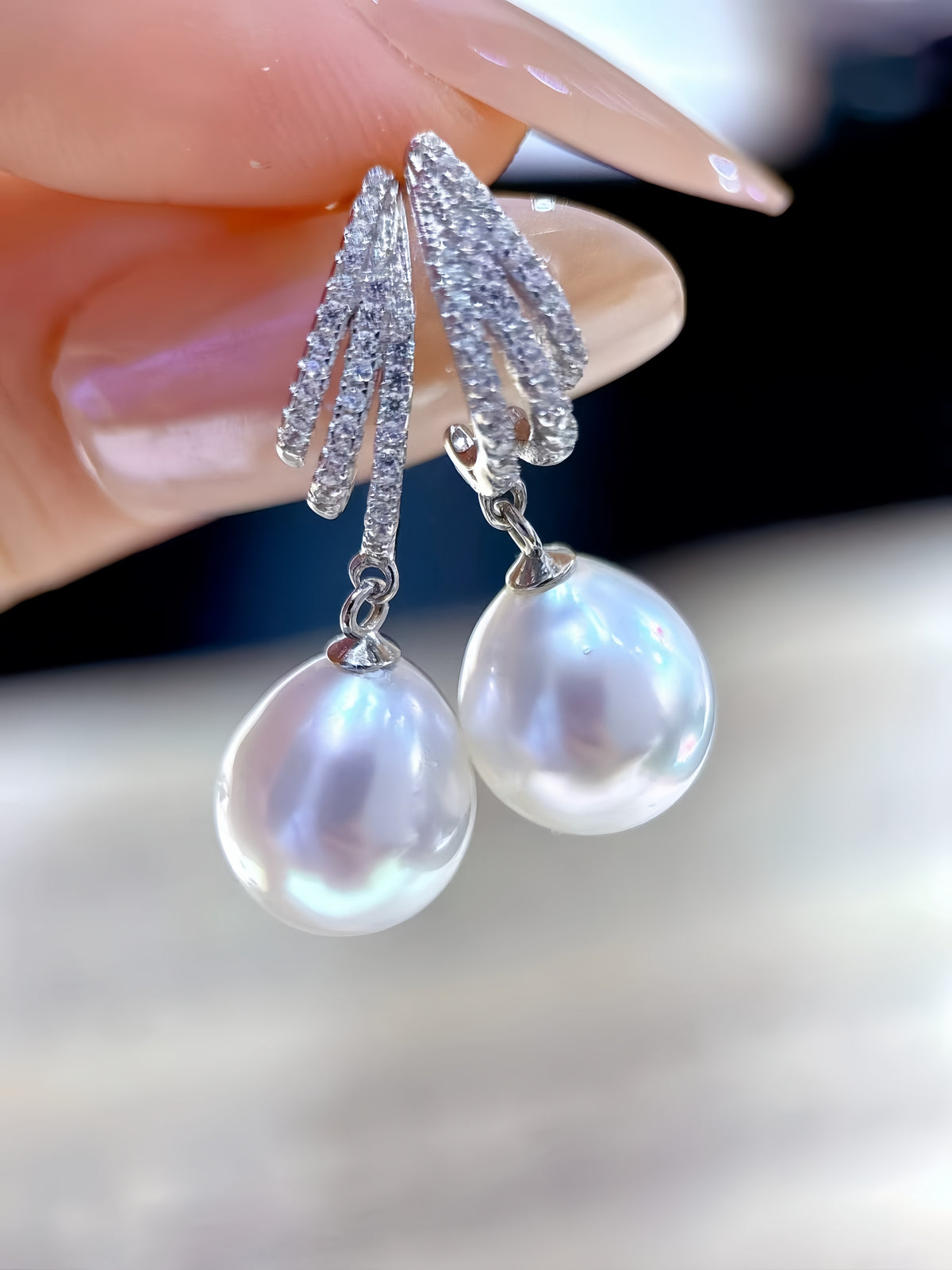 A close-up of a hand holding a pair of silver dangle earrings with pavé-set clear stones and large white pearl drops against a blurred background.
