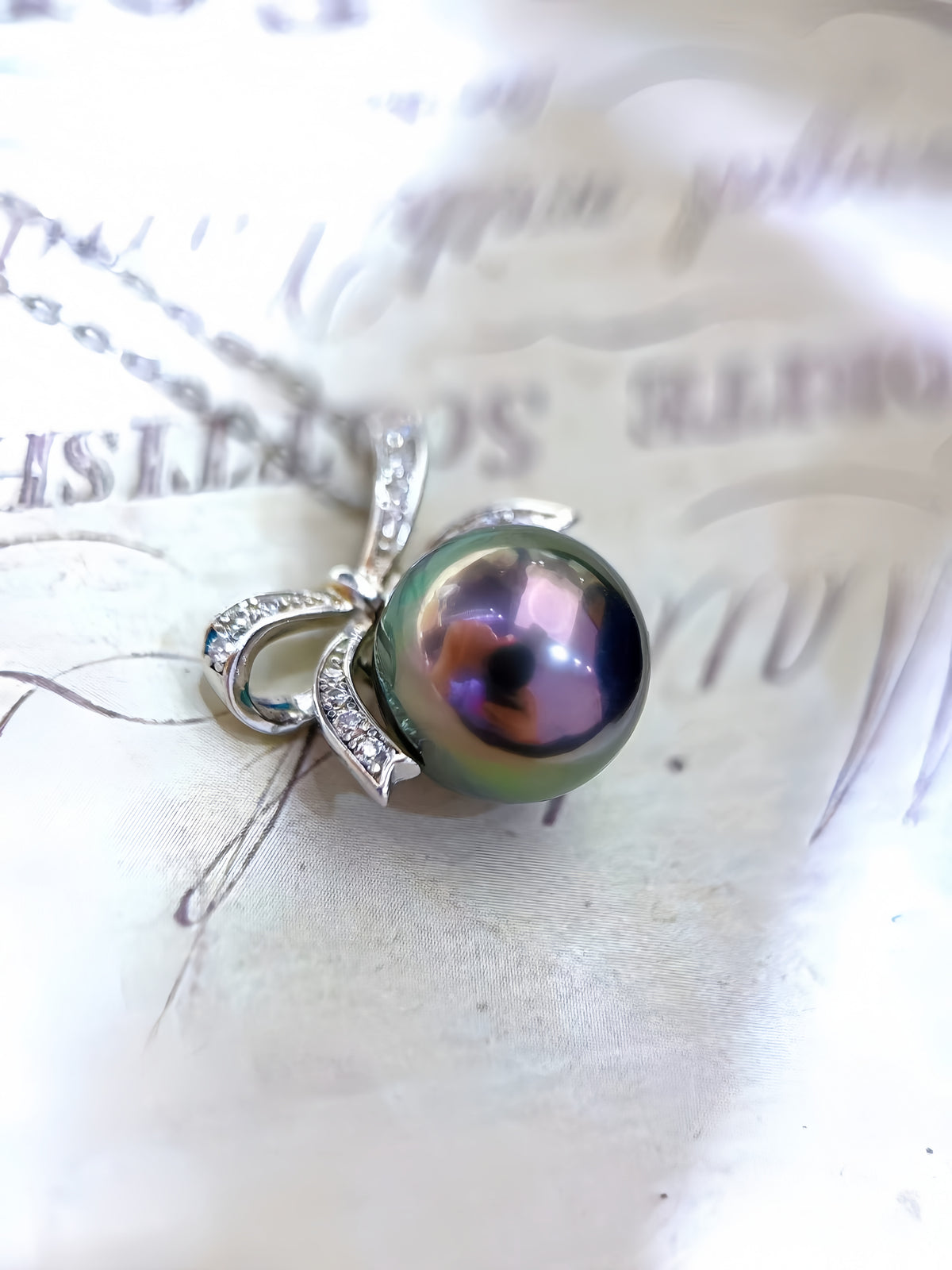 A close-up of a silver pendant necklace featuring a round, iridescent green-black pearl with purple reflections, set in a silver bow-shaped frame with small clear stones, resting on a light-colored textured surface.