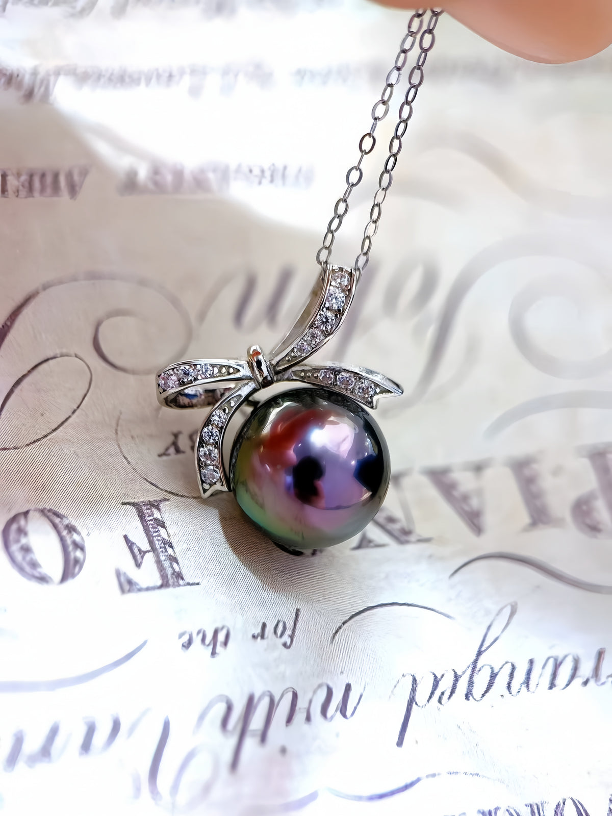 A close-up of a silver pendant necklace featuring a large, iridescent green-black pearl nestled within a silver bow-shaped setting adorned with small clear stones, resting on a decorative cream-colored paper background.