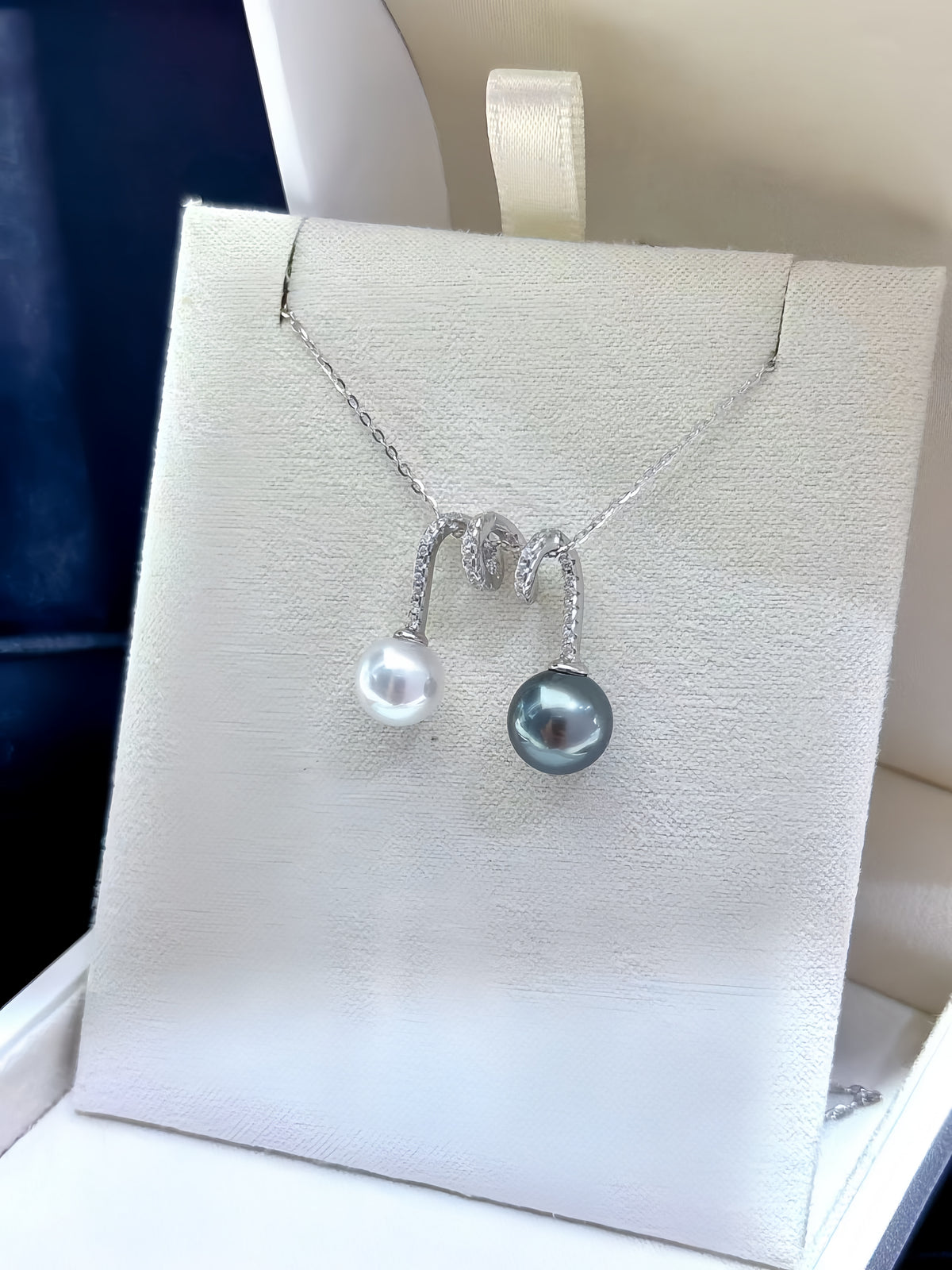A close-up of a silver pendant necklace held on a finger. The necklace features a curved silver chain with small clear stones and two pearl drops: one white and one dark gray/black Tahitian pearl.