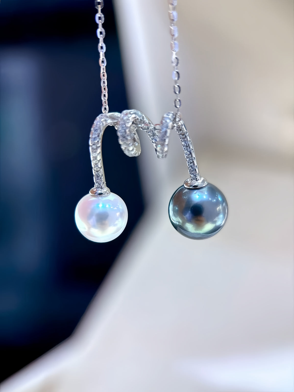 A close-up of a silver pendant necklace held on a finger. The necklace features a curved silver chain with small clear stones and two pearl drops: one white and one dark gray/black Tahitian pearl.