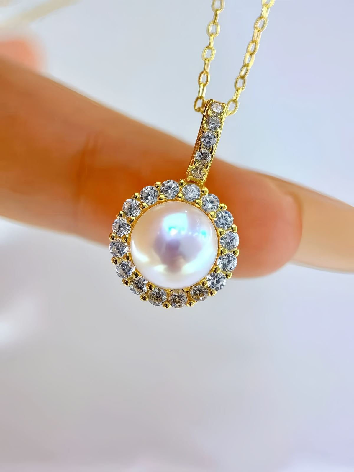 A gold pendant necklace with a central iridescent pearl surrounded by small clear stones, held between fingers against a soft white background.