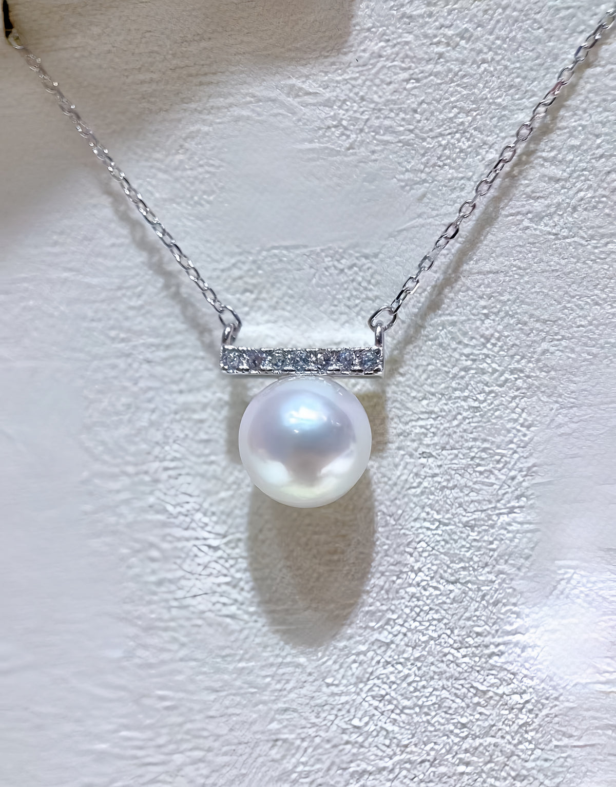 A silver bar necklace featuring a single lustrous white pearl pendant and a small row of clear stones on the bar, displayed against a textured white background.