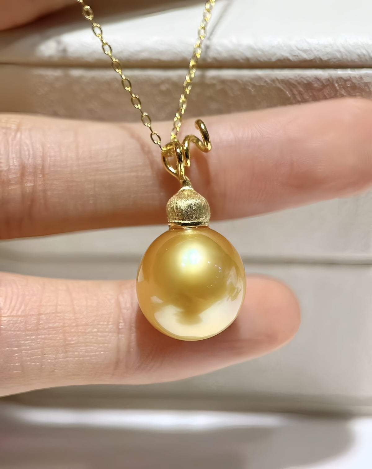 A gold pendant necklace featuring a large, lustrous yellow pearl pendant held by a person's fingers against a light-colored background.