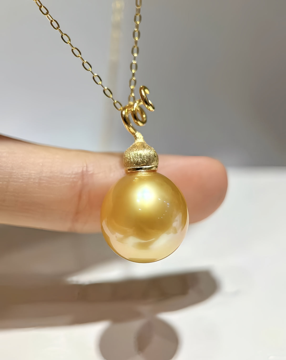 A gold pendant necklace featuring a large, lustrous golden pearl suspended from a delicate gold chain, held by a person's finger against a neutral light gray background.