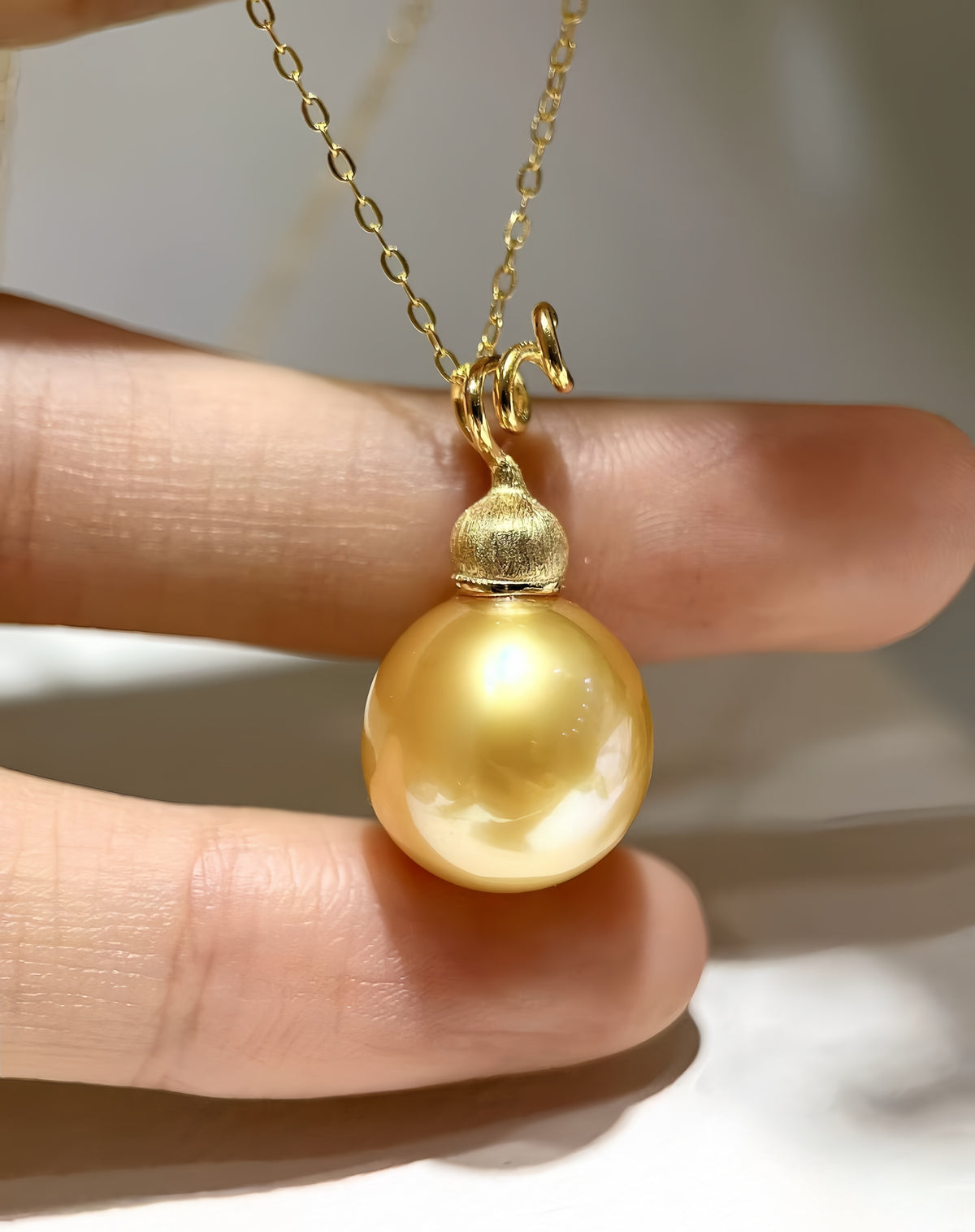 A close-up of a gold pendant necklace featuring a large, lustrous yellow pearl pendant held between fingers.