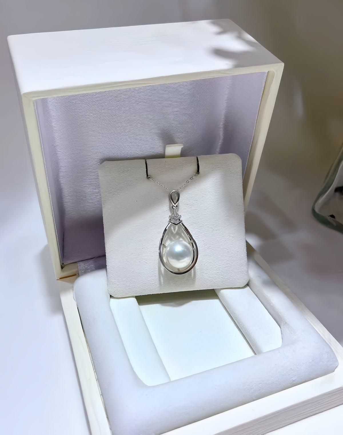 A teardrop-shaped pearl pendant in a silver-toned setting, displayed on a white jewelry card inside an open cream-colored presentation box.