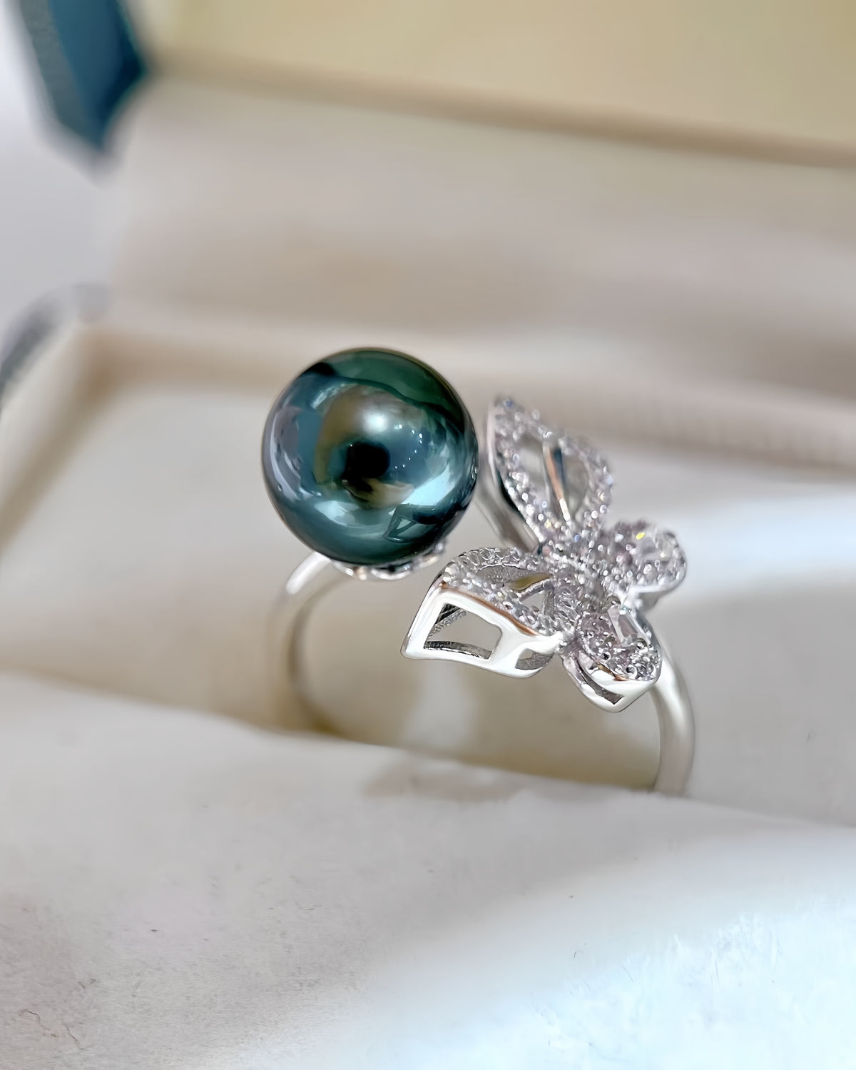 A silver ring featuring a large round dark green pearl and a butterfly-shaped setting paved with small clear stones, displayed in a cream-colored jewelry box.
