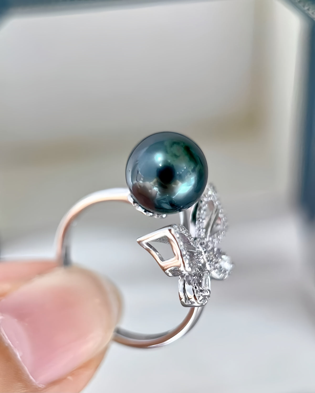 A close-up of a silver ring held by a finger, featuring a round dark green-black pearl and a butterfly-shaped setting with small clear stones, against a soft light gray background.