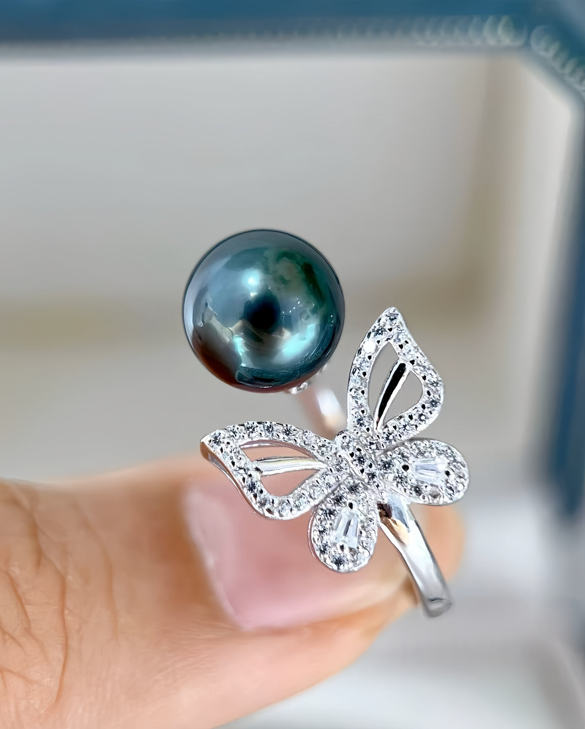 A close-up of a silver butterfly ring with clear pavé stones and a dark teal freshwater pearl on a finger.