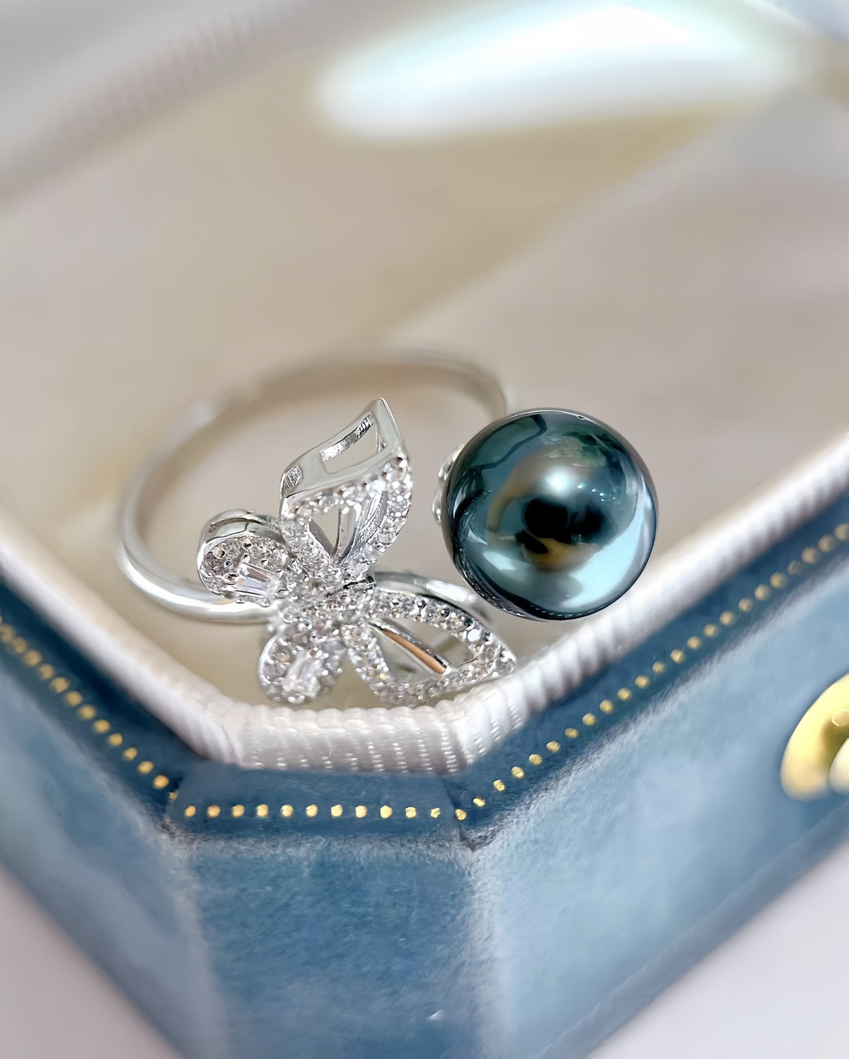A silver ring featuring a butterfly motif set with small clear crystals and a large, dark blue-gray pearl, displayed inside a blue ring box.