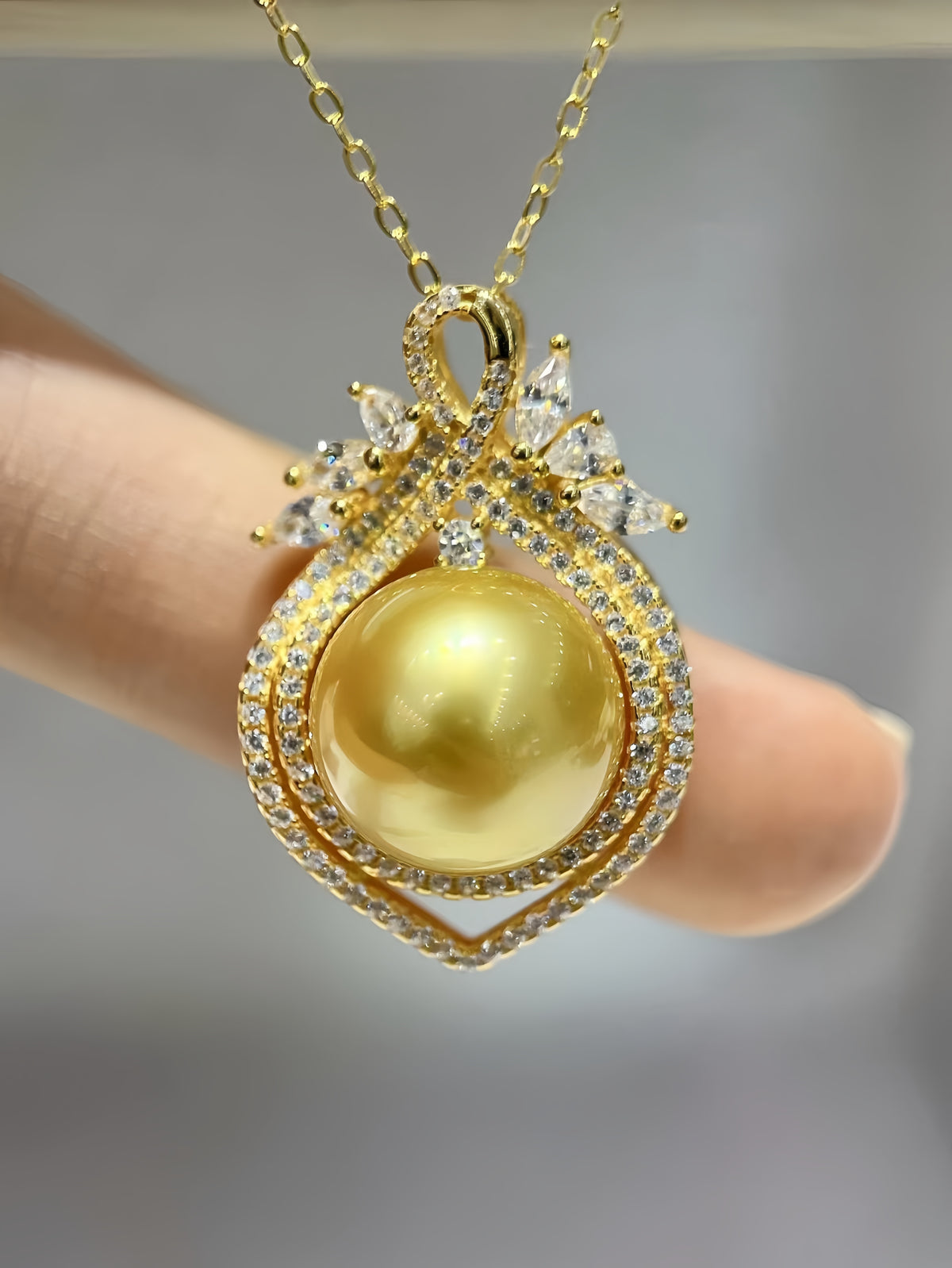A gold pendant necklace with a large round yellow pearl, surrounded by pavé-set clear stones and a bow-like top detail, shown being held against a soft gray background.