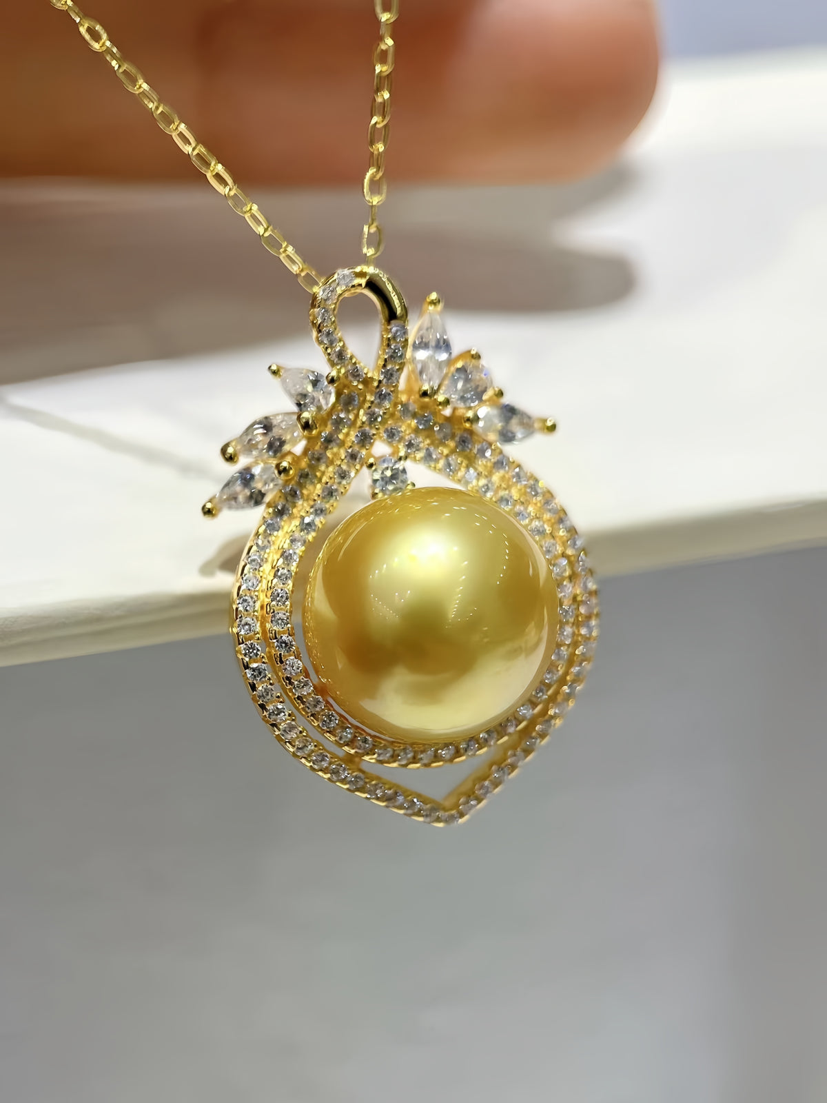 A gold pendant necklace featuring a large round yellow pearl surrounded by clear pavé stones, hanging from a gold chain against a soft white background.