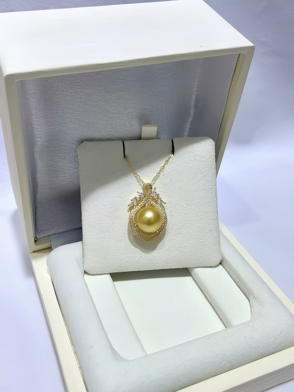 A gold pendant necklace with a large yellow pearl and small clear stones, displayed on a cream jewelry box insert.
