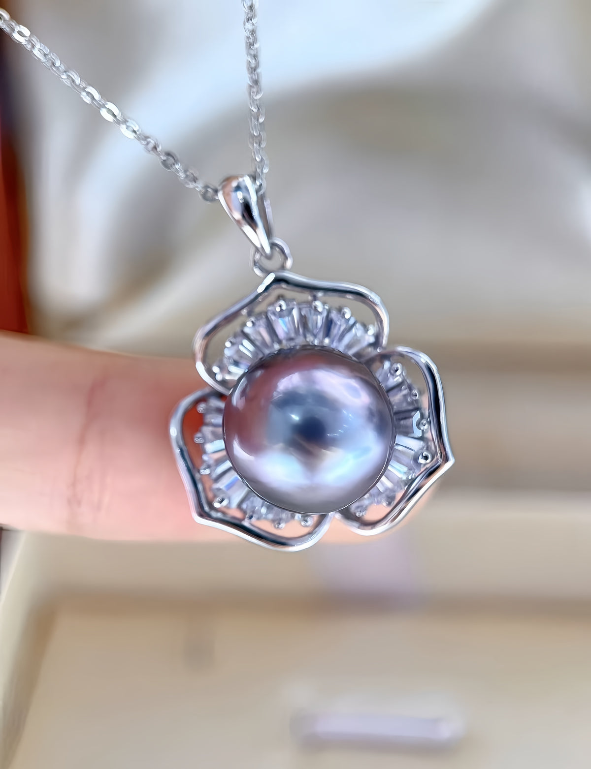 A silver pendant necklace featuring a large round iridescent pearl surrounded by small clear stones in a flower-like setting, held by a person's fingers against a soft beige background.