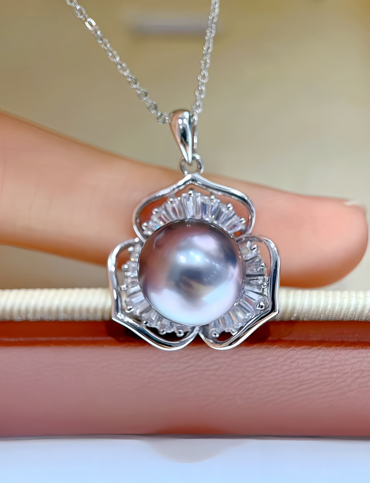 A silver pendant necklace featuring a large lustrous pearl surrounded by clear baguette and round stones, held against a soft beige background with a finger visible behind it.