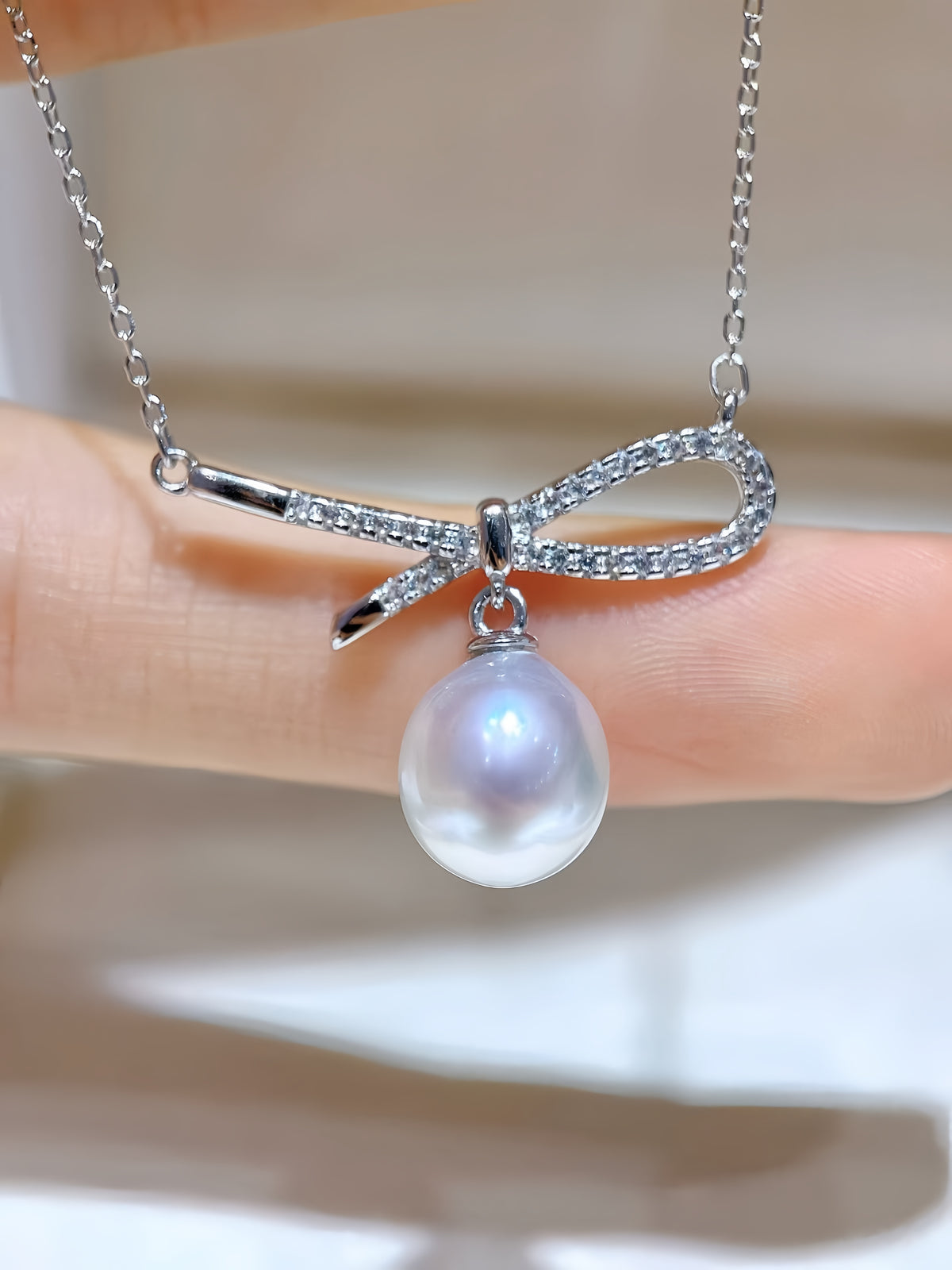 A silver bow pendant necklace with a single lustrous pearl hanging below, held against a soft beige background.