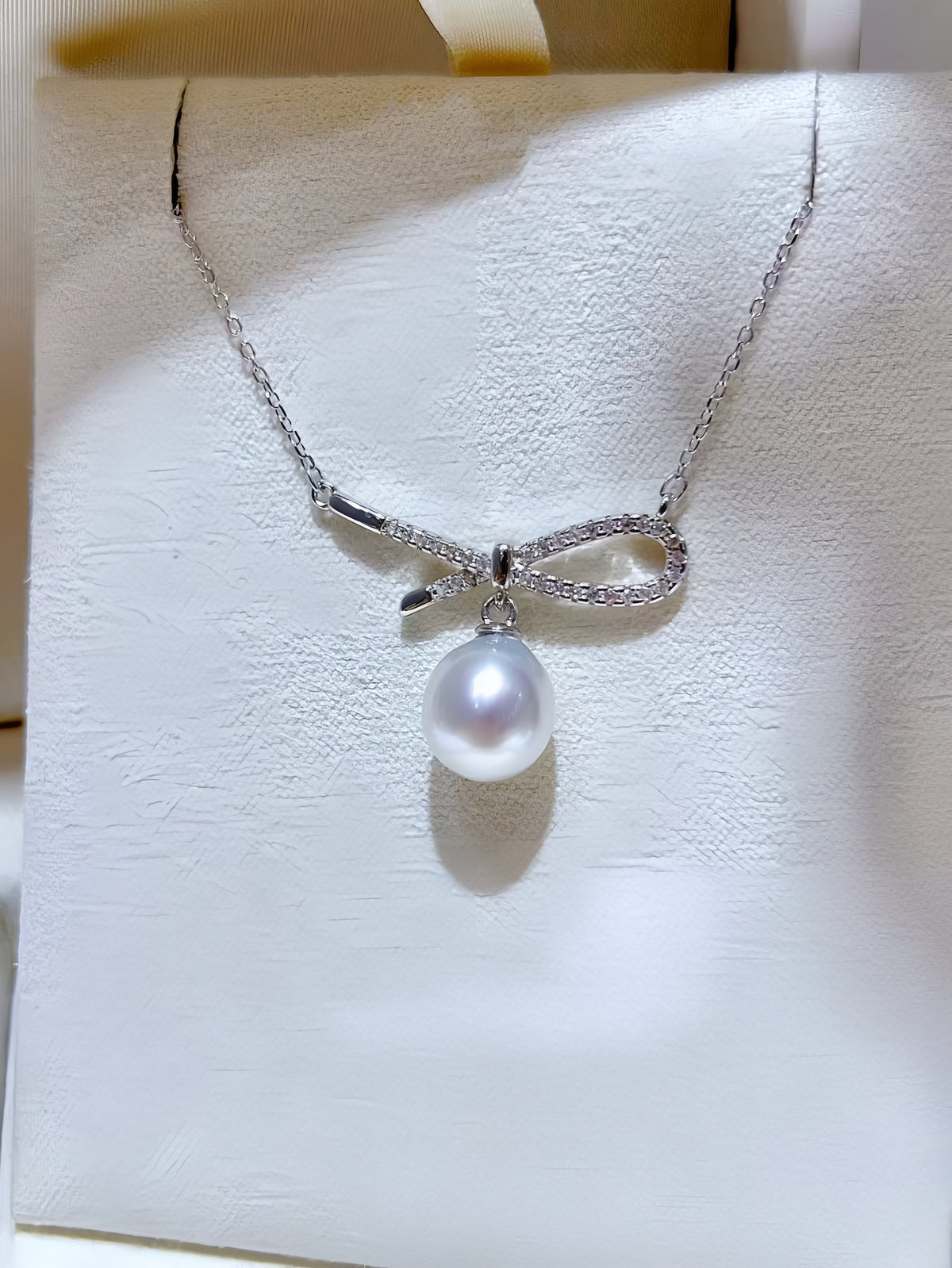 A silver bow pendant necklace with a single lustrous pearl hanging below, held against a soft beige background.