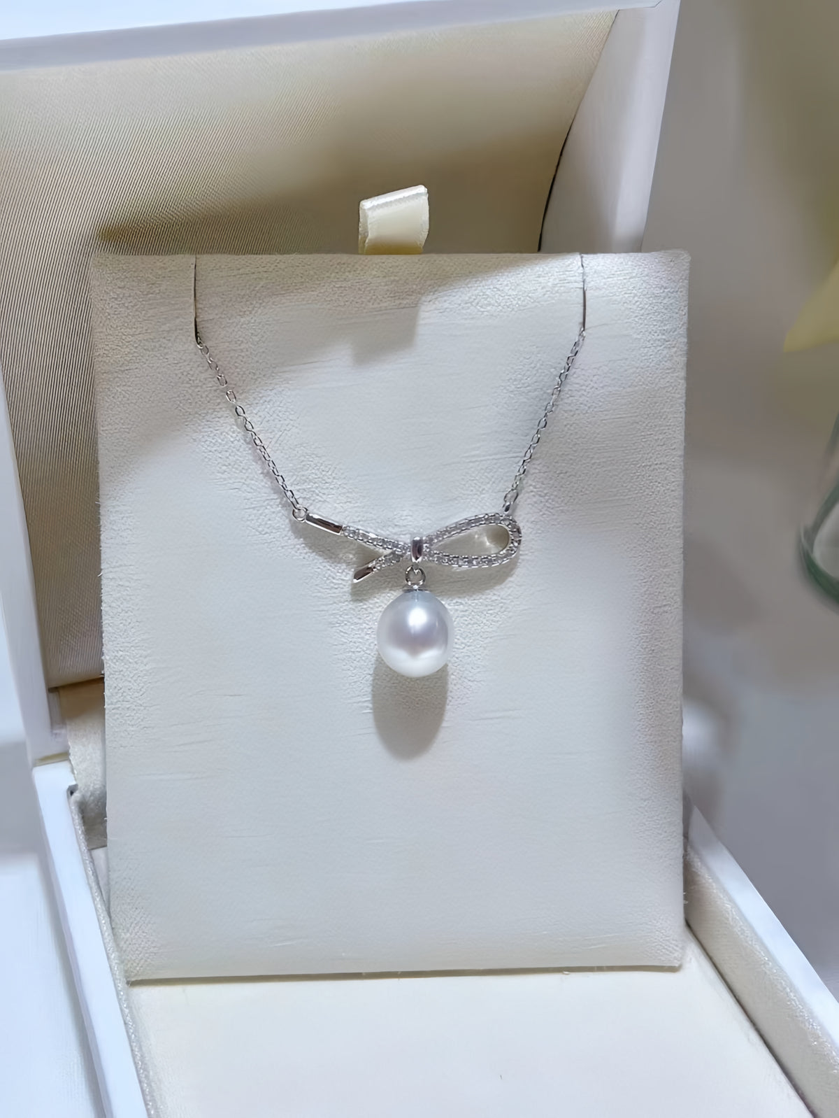 A silver bow pendant necklace with a single lustrous pearl hanging below, held against a soft beige background.