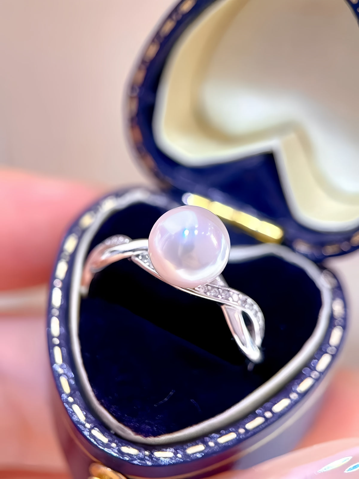 A silver ring with a round white pearl and small clear stones, displayed in an open dark blue heart-shaped jewelry box held in a hand.