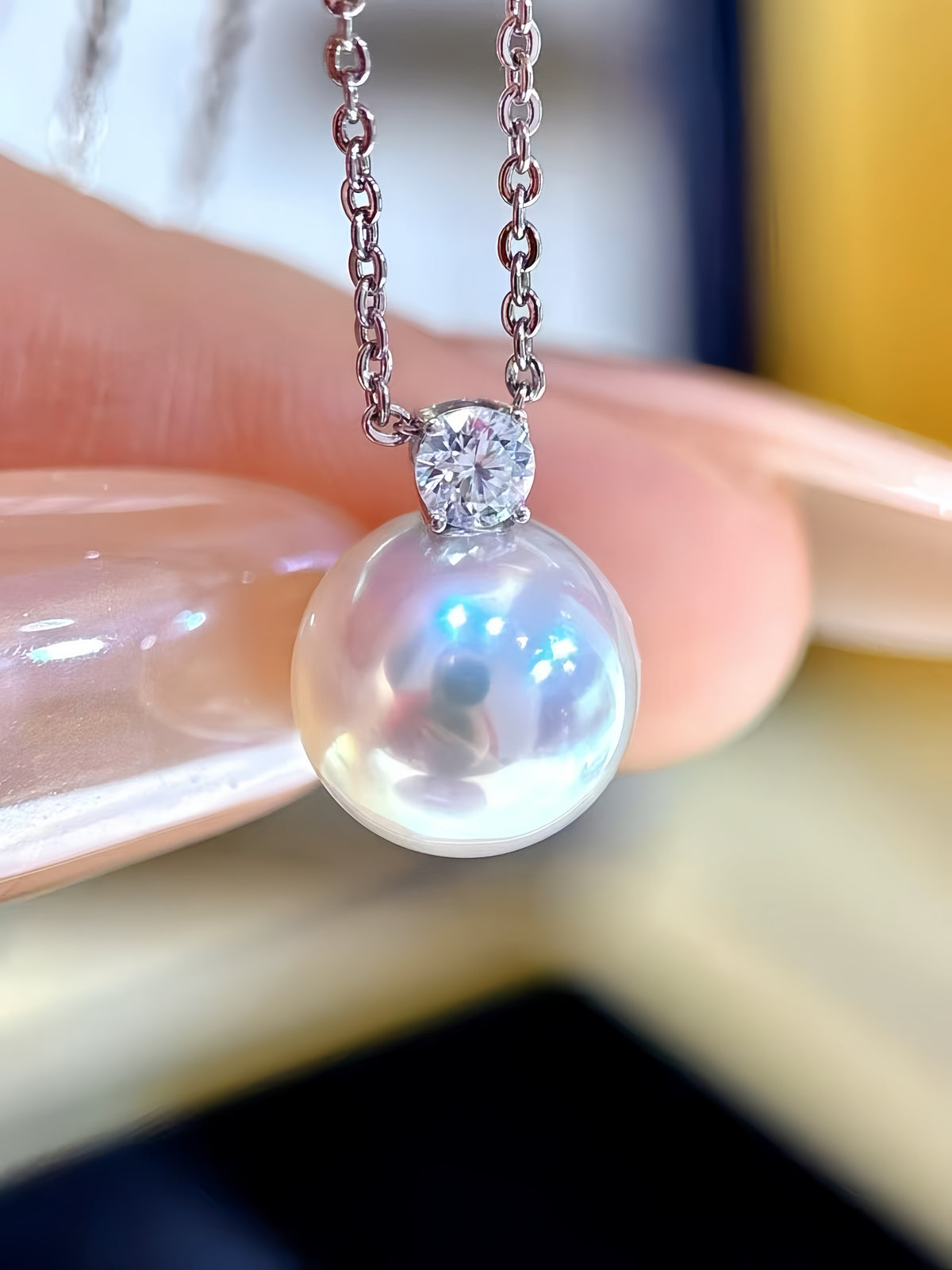 A close-up of a pearl pendant necklace with a silver-toned chain and bezel setting, held between fingers against a softly blurred background.