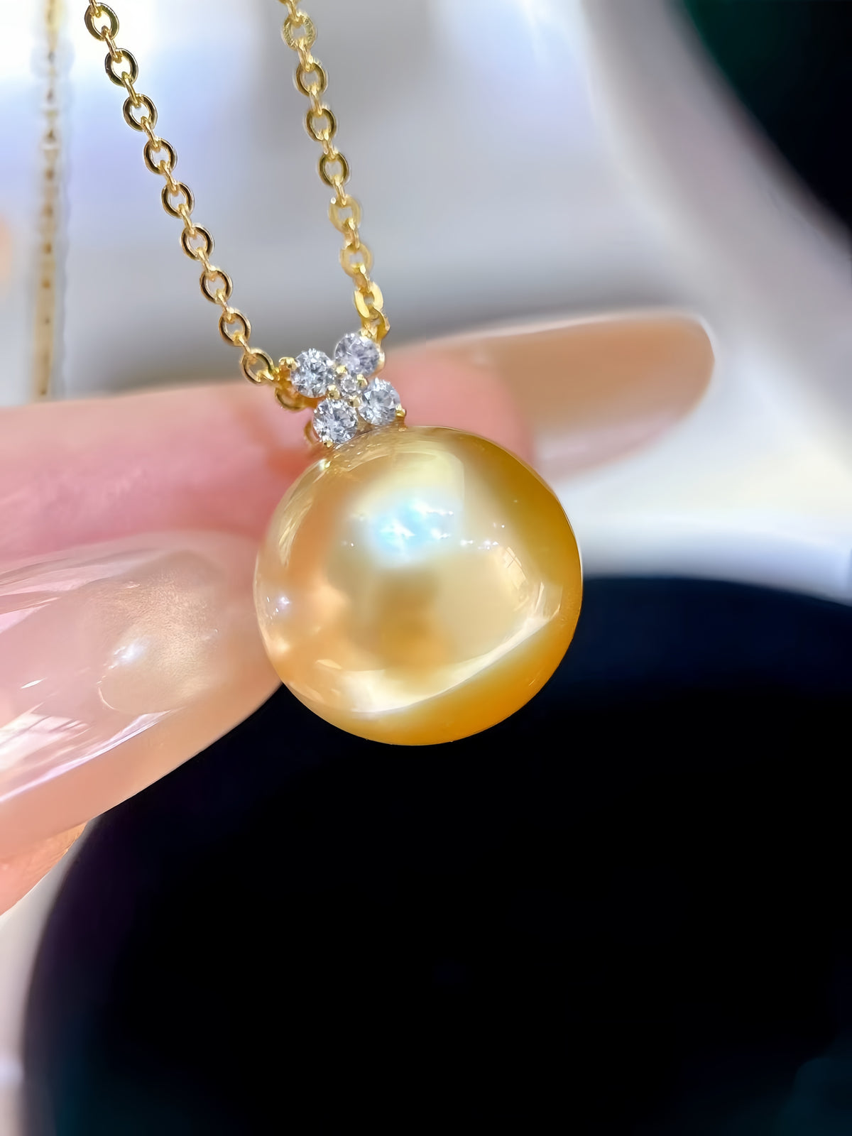 A close-up of a gold chain necklace with a large, round, lustrous golden pearl pendant held by a hand. The pendant is attached to a small, clear, diamond-like floral cluster at the top.