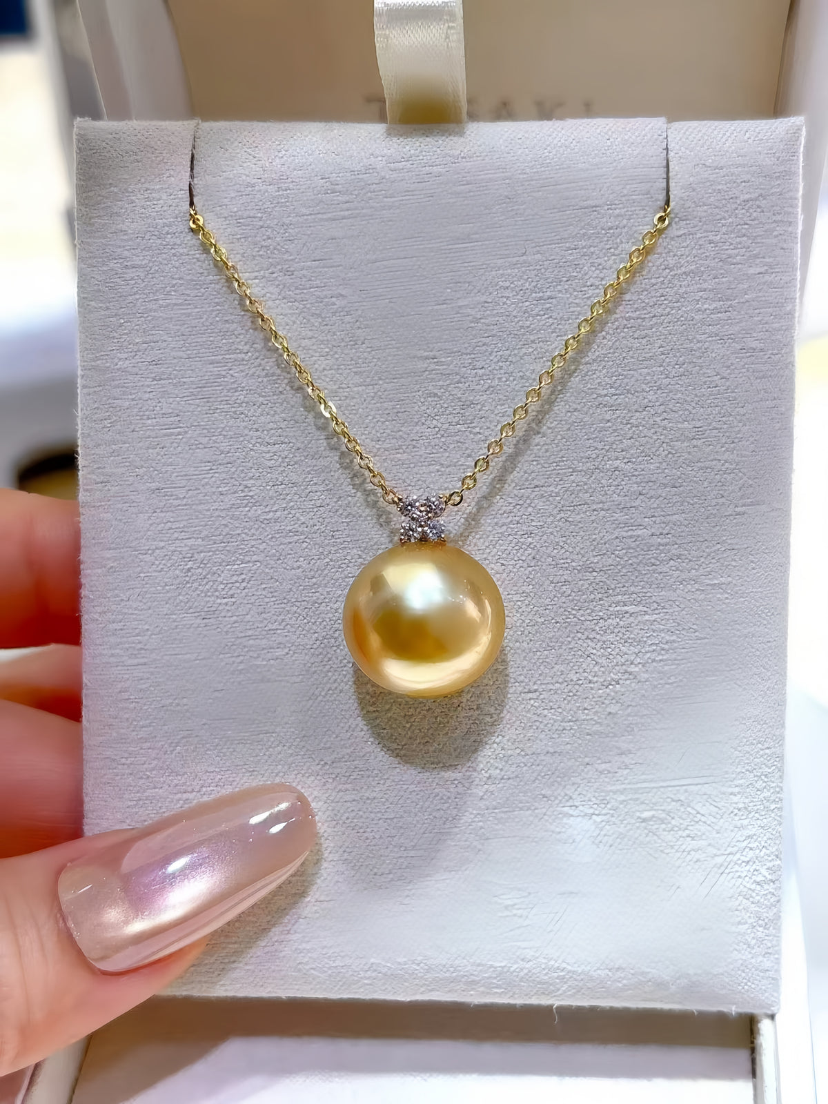 A gold necklace with a large round yellow pearl pendant displayed on a white jewelry card, held by a hand with iridescent nail polish.