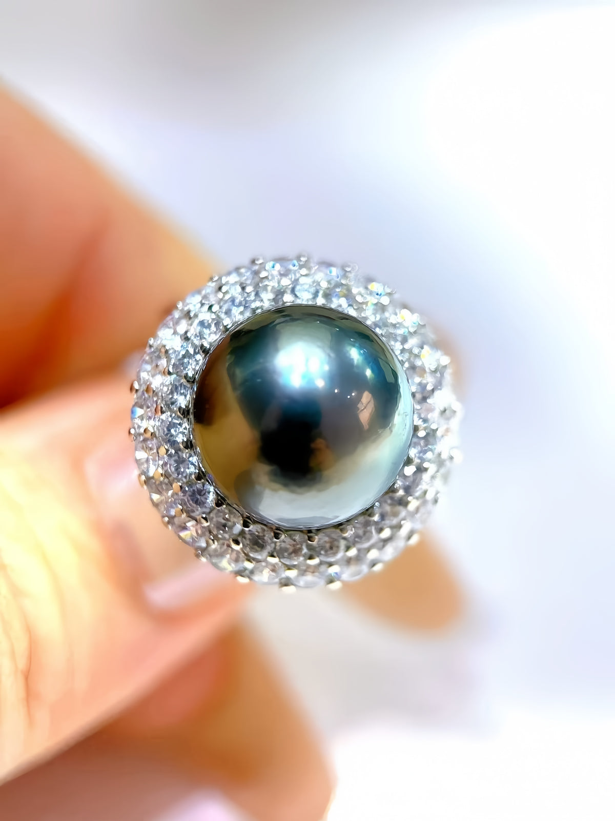 A close-up of a ring held between fingers, featuring a large round dark gray pearl surrounded by a halo of small clear stones on a white background.
