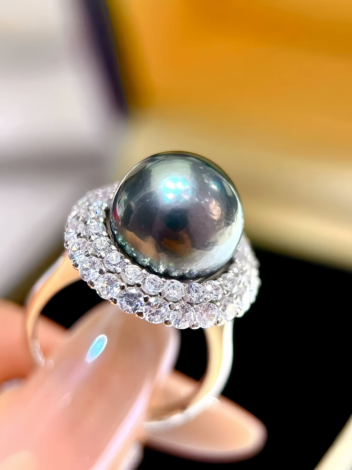 A close-up of a ring being held by a hand, featuring a large round dark gray pearl surrounded by a halo of small clear stones on a gold band.