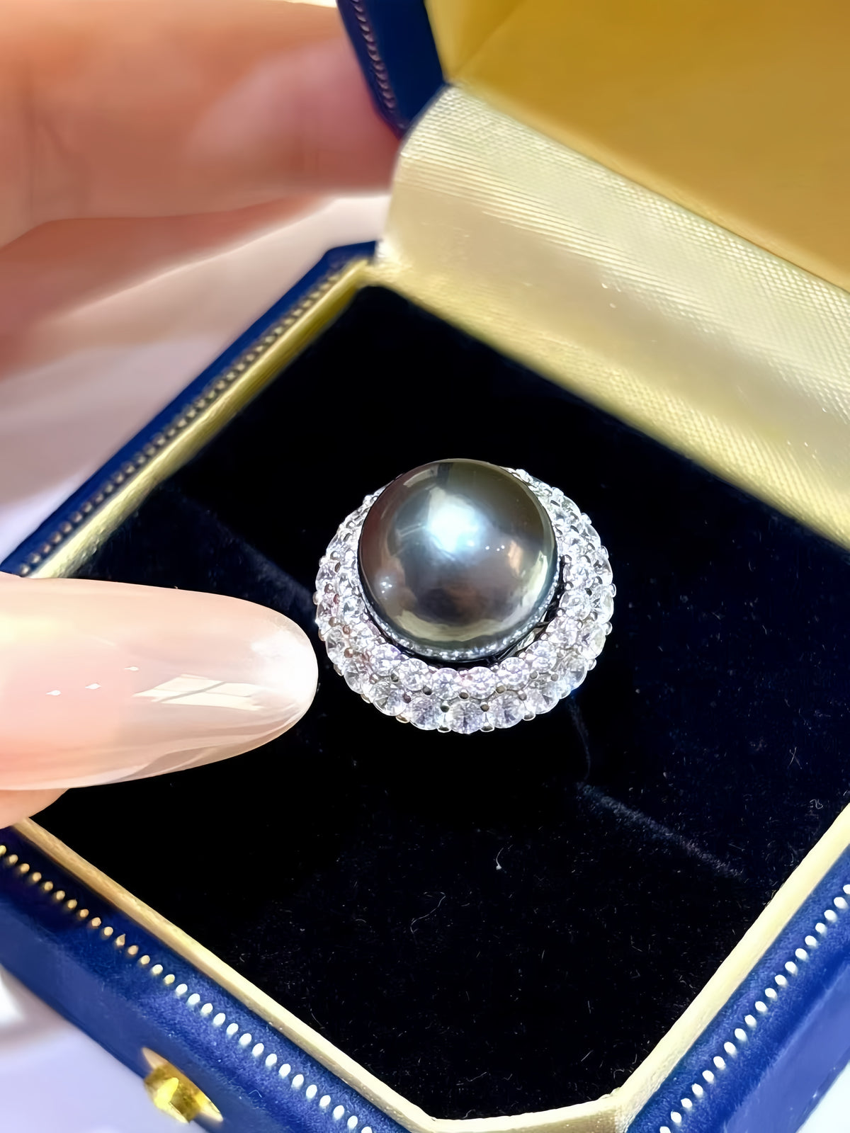 A close-up of a ring with a large dark pearl surrounded by a halo of small clear stones, displayed in a blue ring box with a gold interior. A hand is holding the box open.