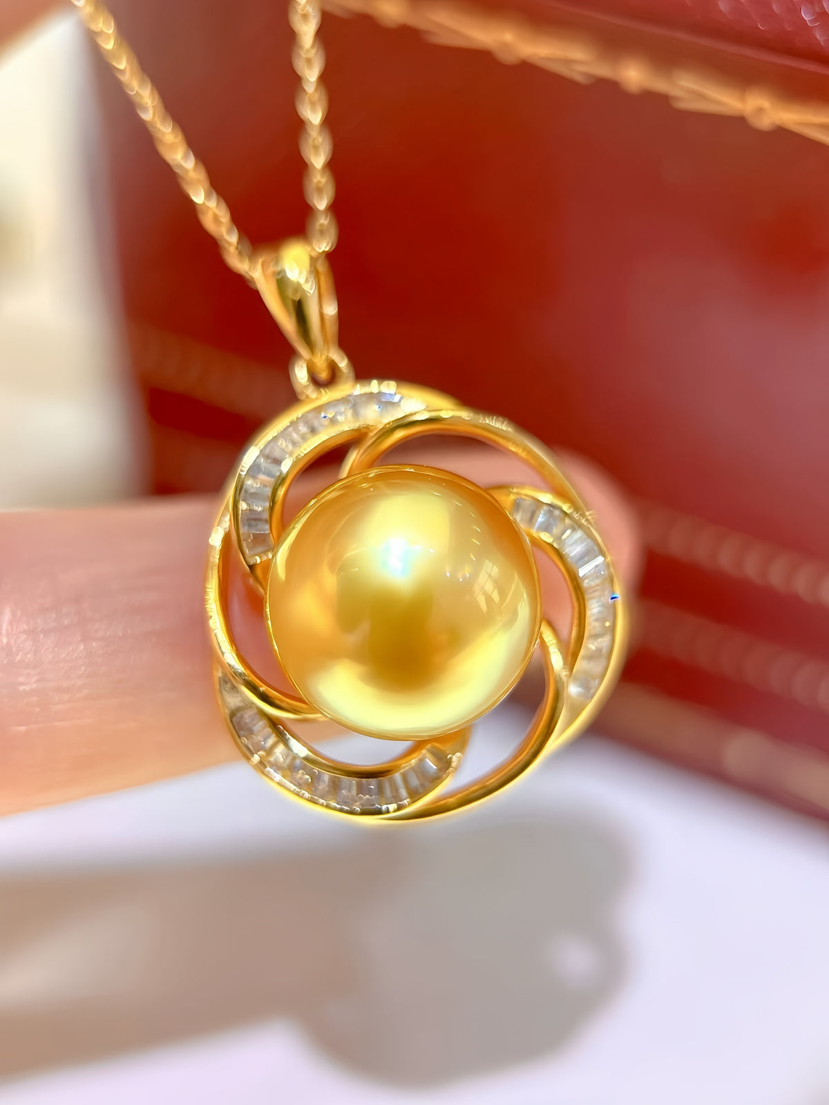 A gold pendant necklace featuring a large round yellow pearl in the center, surrounded by open swirls of gold metal set with small clear stones, held near a finger against a blurred red and white background.