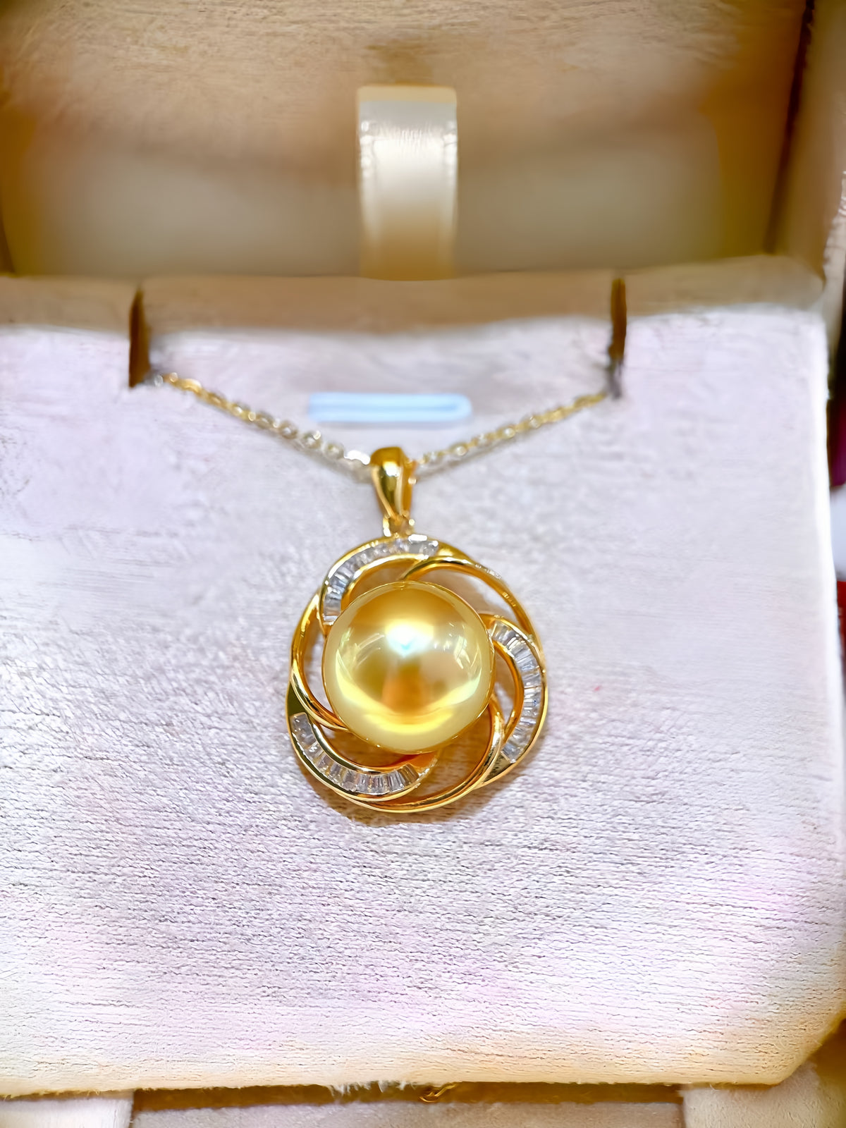 A gold pendant necklace featuring a large round yellow pearl surrounded by swirling gold and small clear stones, displayed inside a cream-colored jewelry box.