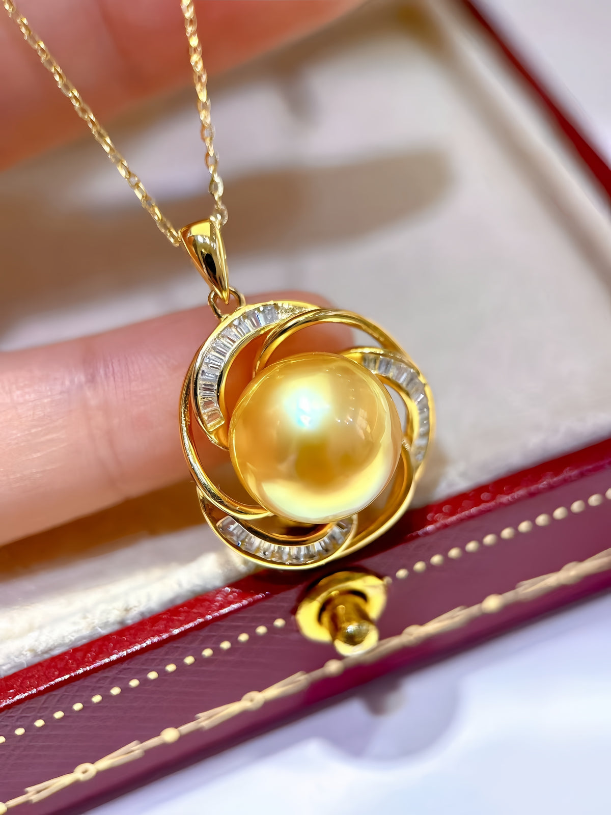 A gold pendant necklace featuring a large, lustrous yellow pearl surrounded by three interlocking gold rings, one of which is set with small clear stones. The necklace is held by a hand over a red jewelry box.