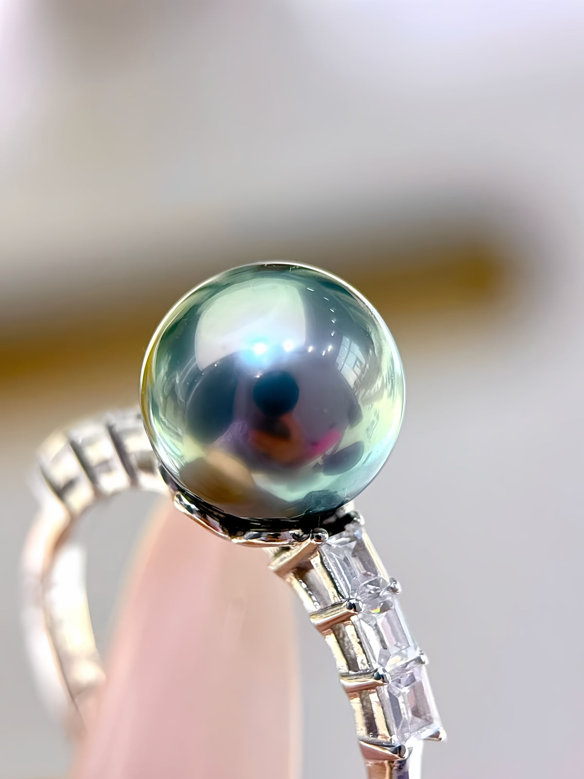 A close-up of a silver ring featuring a large, round, iridescent greenish-blue pearl on a bamboo-style setting, held against a soft, blurred background.
