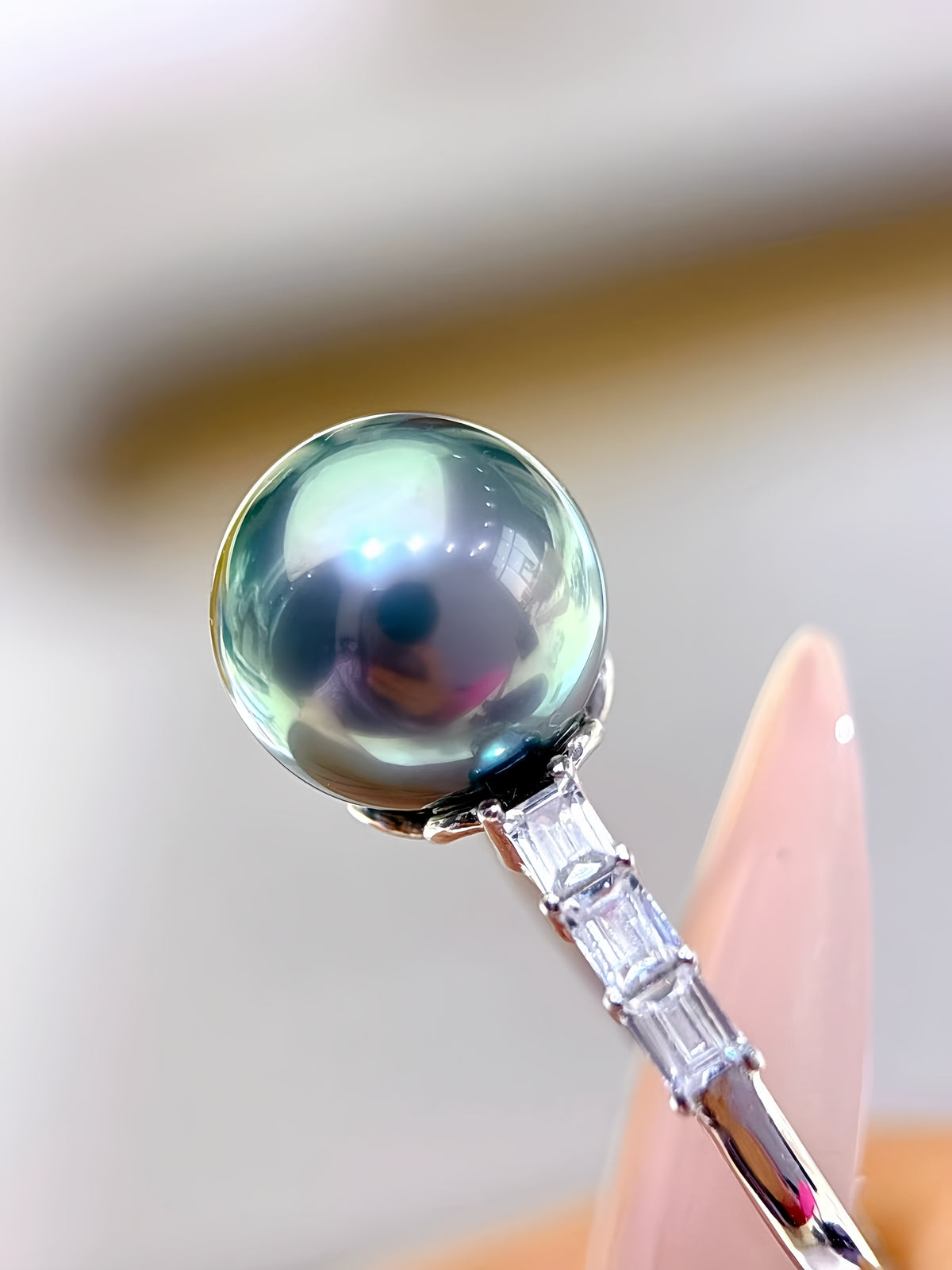 A close-up of a silver ring featuring a large, round, iridescent green pearl and a channel-set band of clear baguette and round crystals, held by a hand with a clean, blurred background.