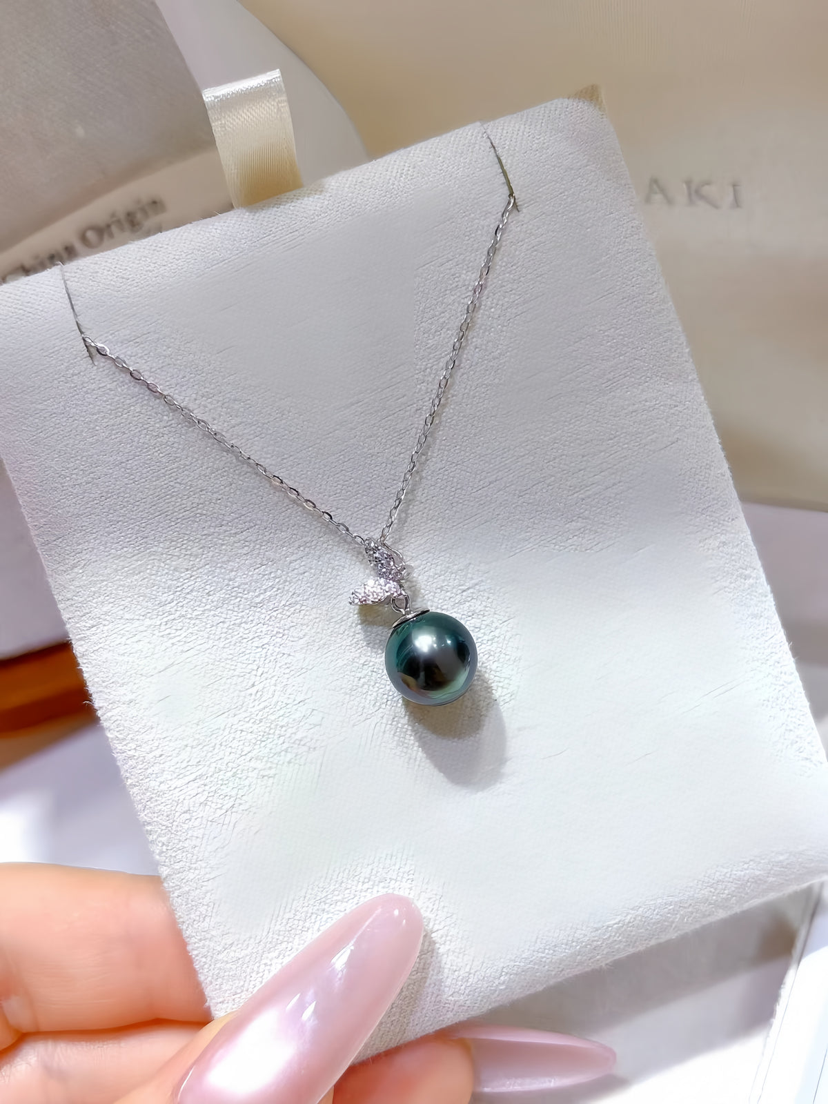 A silver necklace with a dark teal-gray round pearl pendant and a small clear stone, displayed on a white jewelry card held by a hand with pale pink manicured nails.