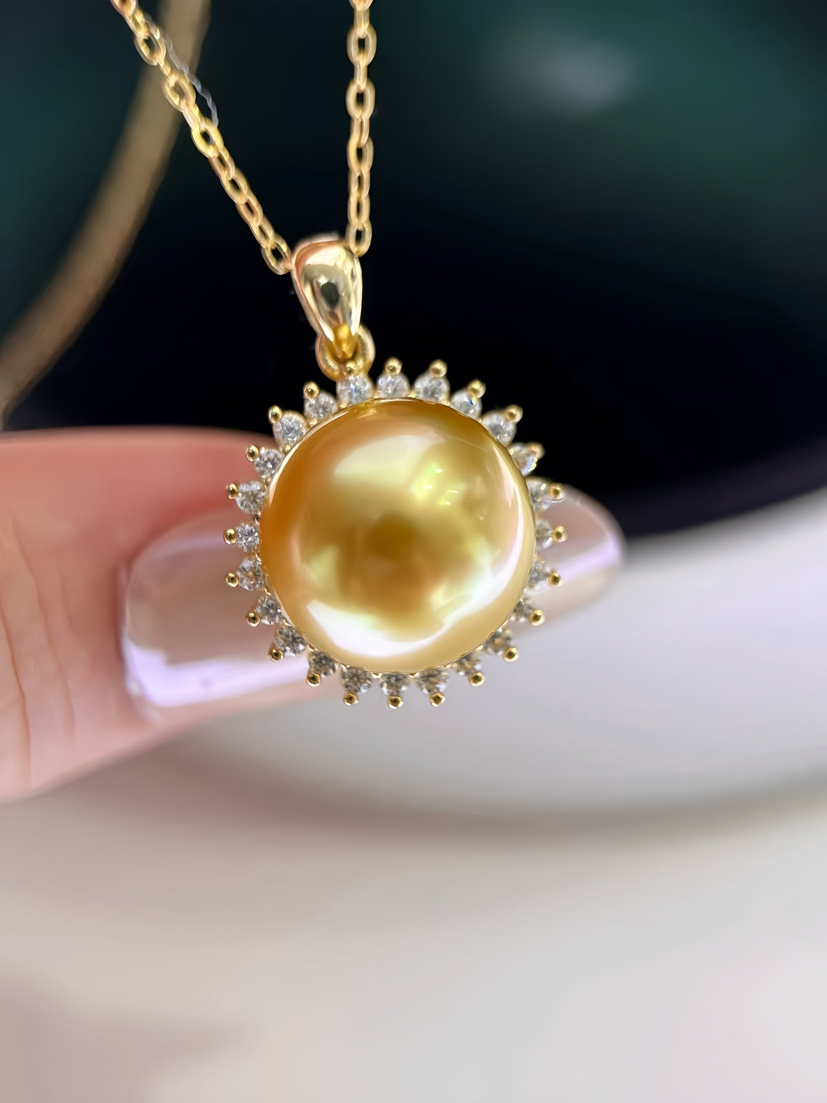 A gold pendant necklace featuring a large, lustrous yellow pearl surrounded by small clear stones, held by a finger against a soft-focus background.