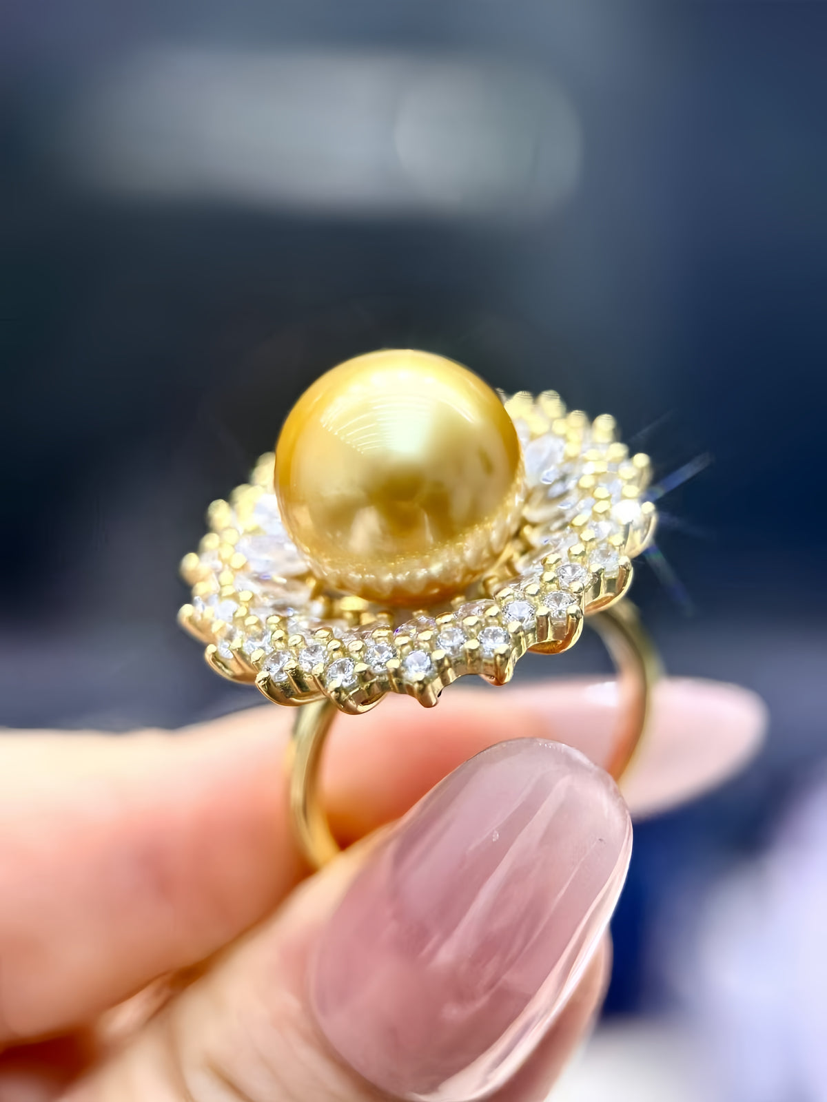 A gold ring featuring a large round yellow pearl in the center, surrounded by a halo of small clear stones, held by a hand with pink manicured nails against a dark blurred background.