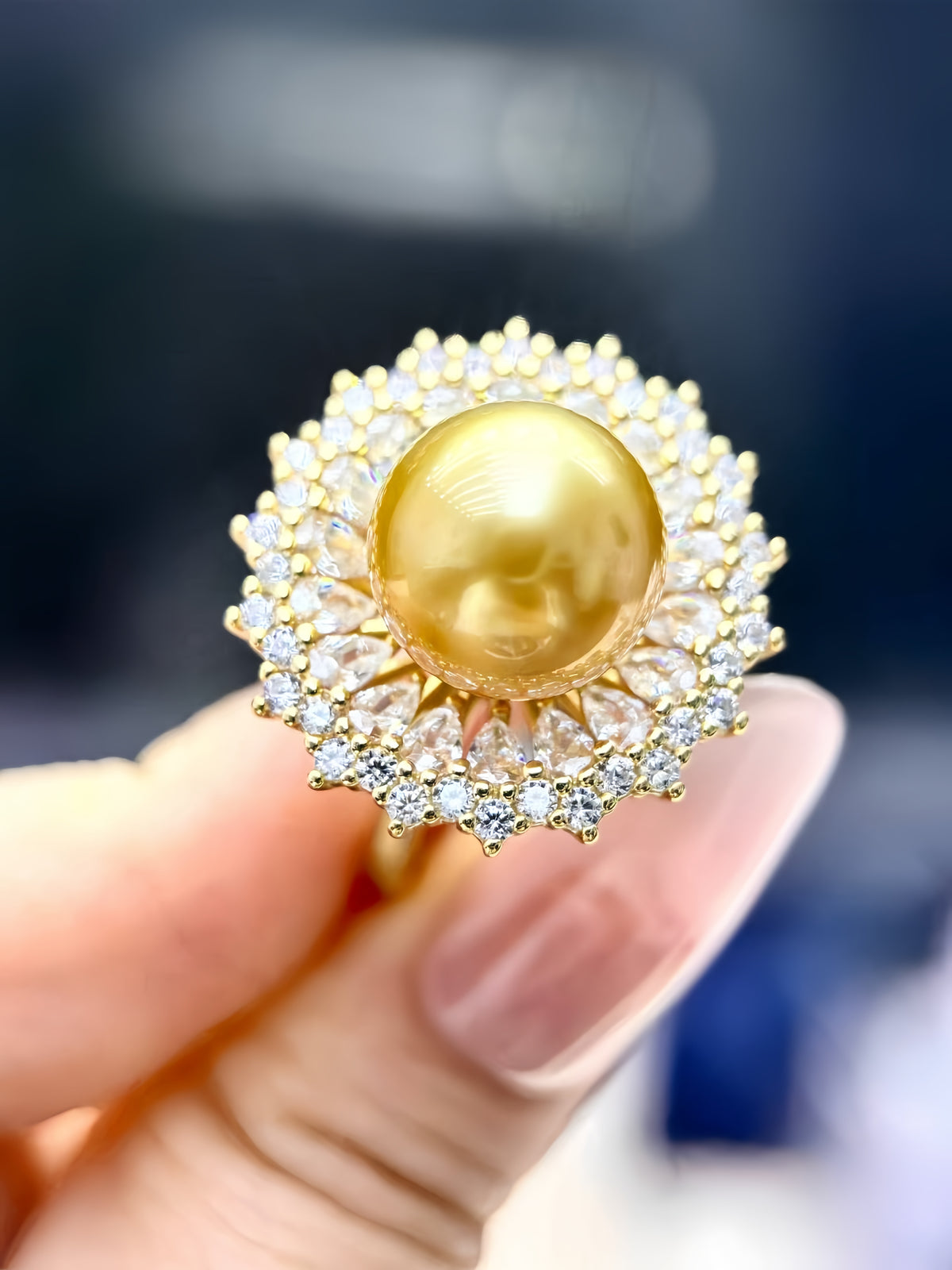 A gold ring featuring a large round yellow pearl surrounded by a halo of small clear stones, held between fingers against a blurred dark background.