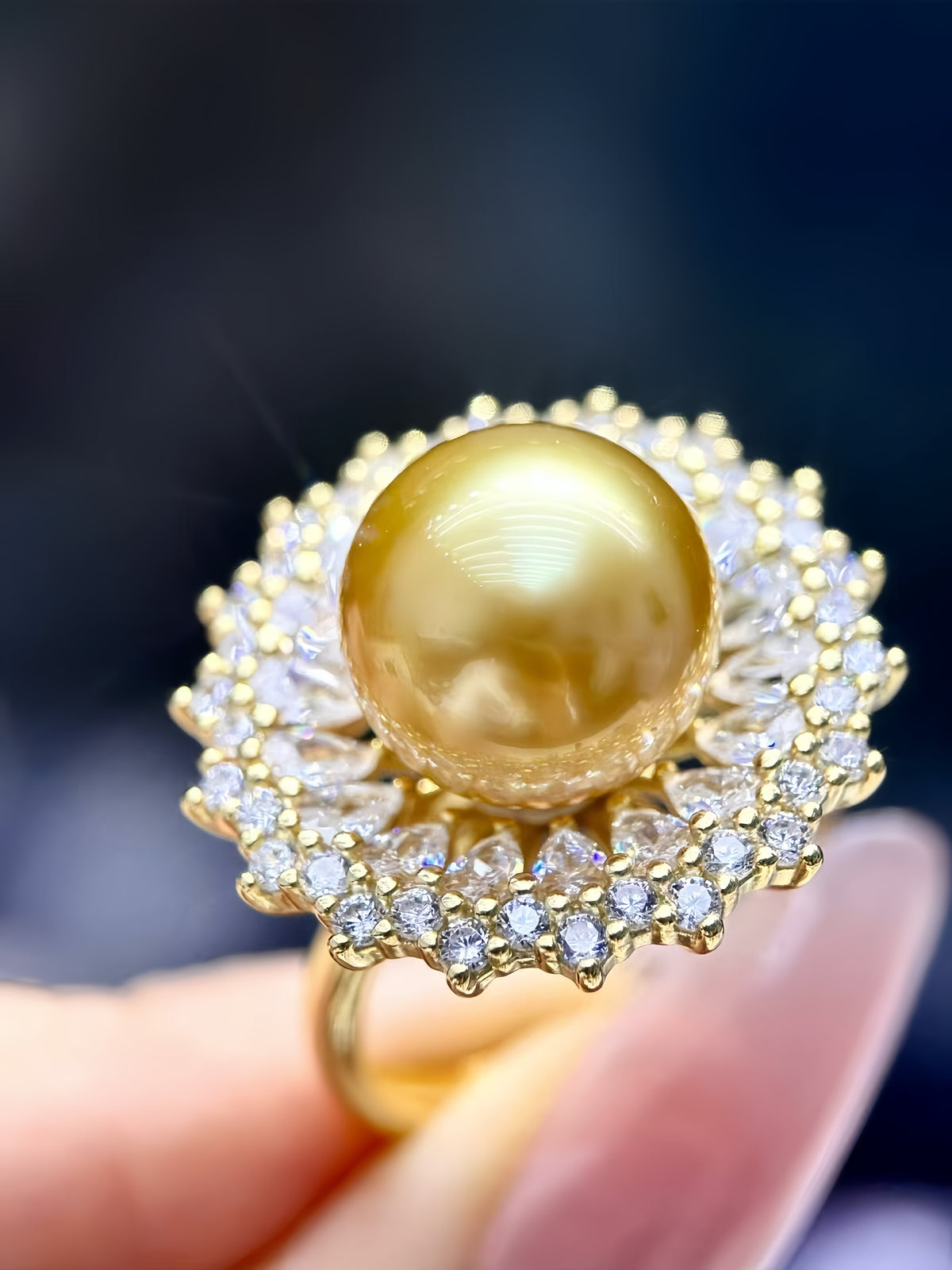 A close-up of a gold ring featuring a large, round, golden-yellow pearl surrounded by a halo of small clear stones, held by a hand.