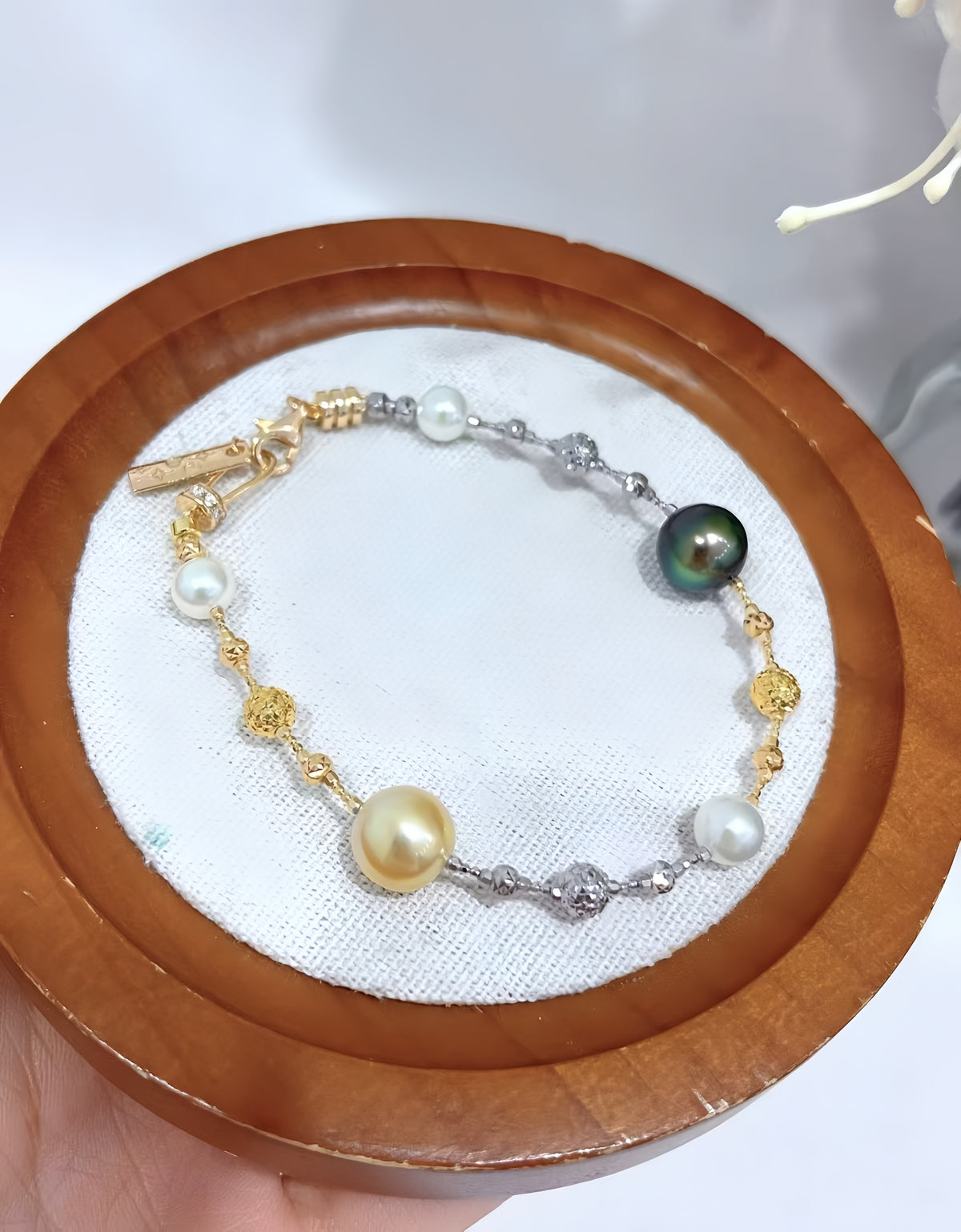 A delicate bracelet with gold and silver-toned links, white pearls, and a dark green teardrop pearl, displayed on a white fabric-lined wooden tray.