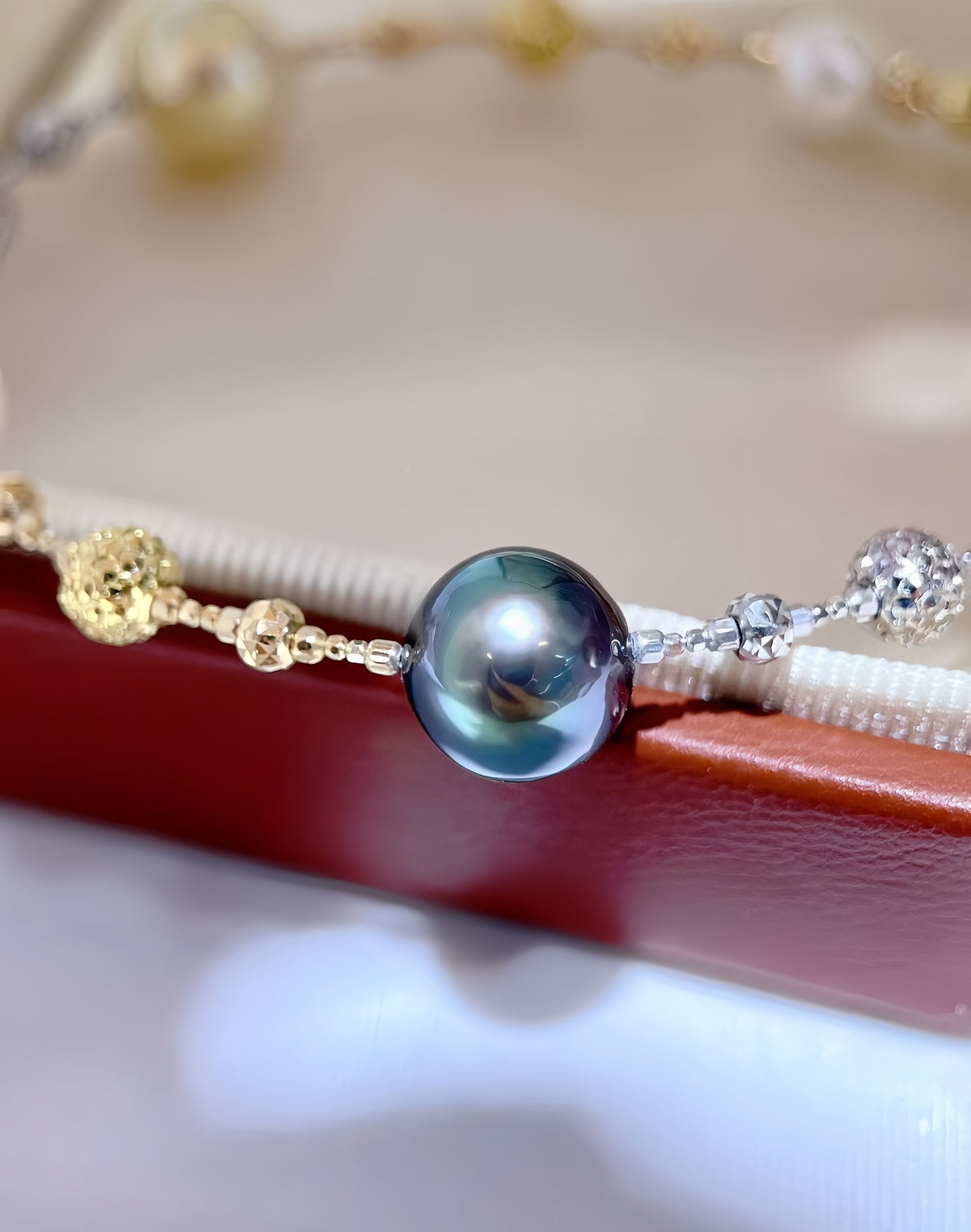 A close-up of a delicate jewelry piece featuring a large, round iridescent dark blue-gray pearl centered on a thin chain with small gold and silver beads, resting on a red and cream display stand.