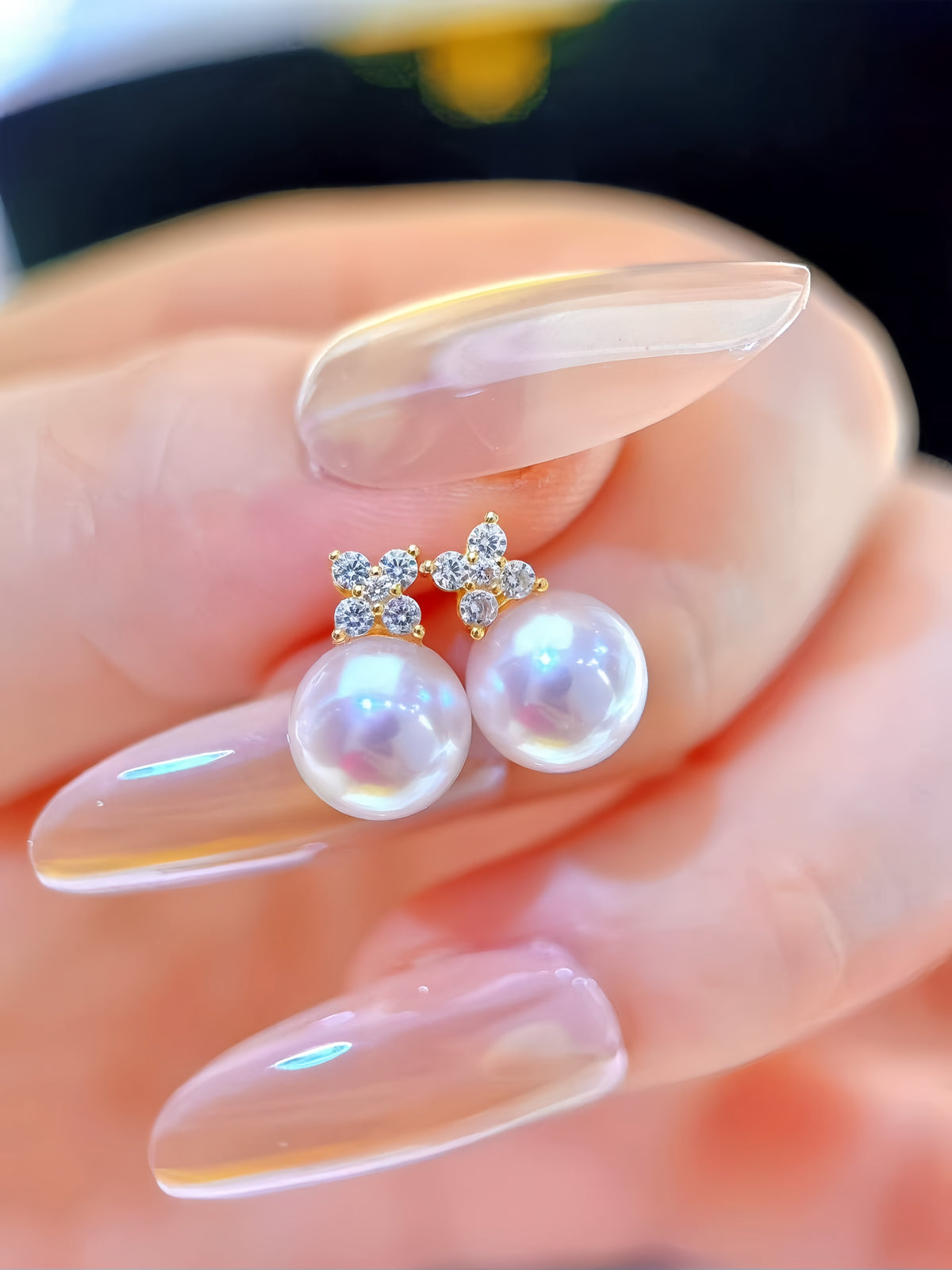 A close-up of a hand with long, glossy nude manicured nails holding a pair of gold bow stud earrings with white pearls and small clear crystal accents.