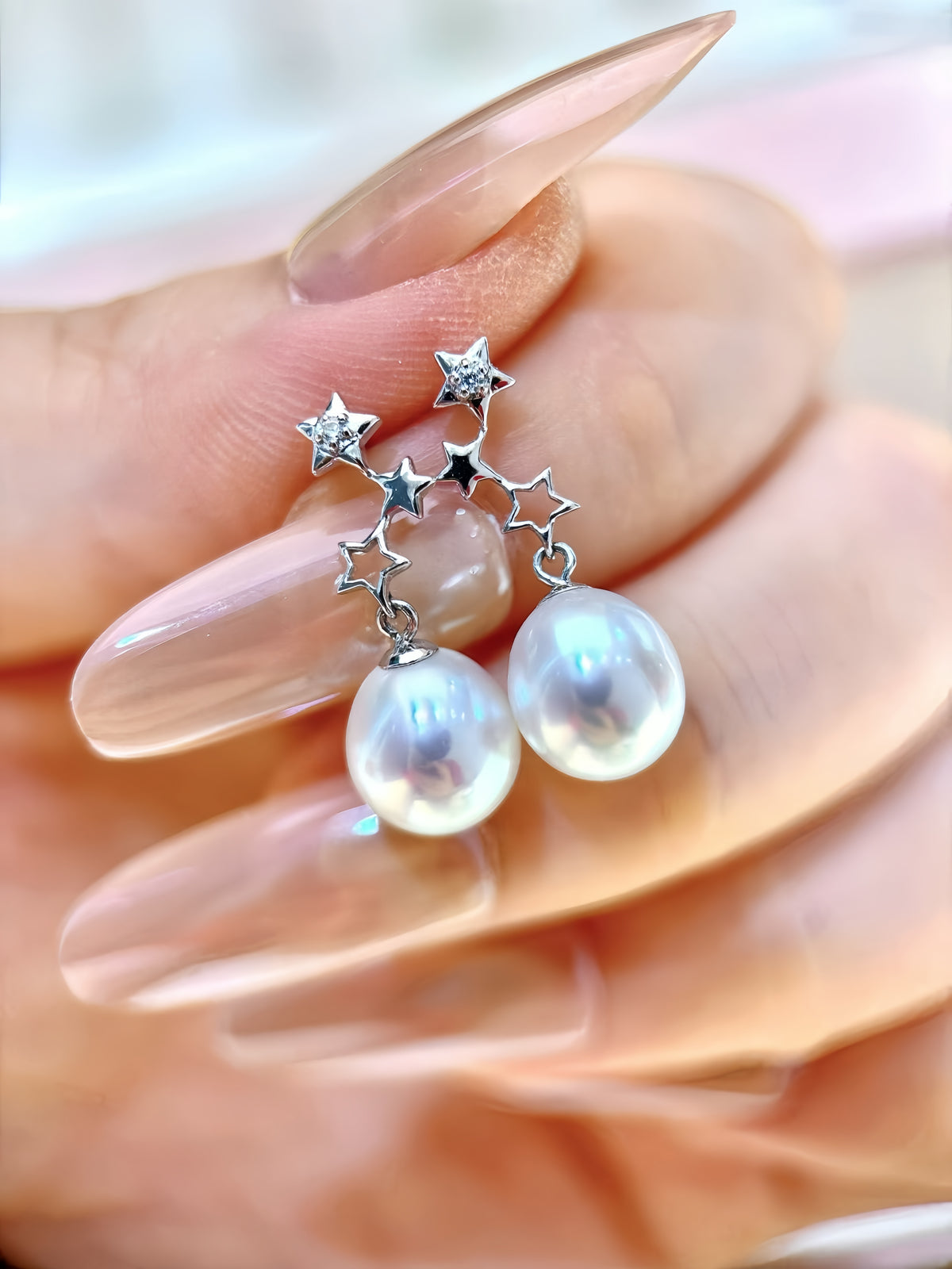 A close-up of a hand with long, glossy nude manicured nails holding a pair of dangling earrings. The earrings feature silver-toned star-shaped charms with small clear stones, connected by a delicate chain to large, lustrous white pearl drops.